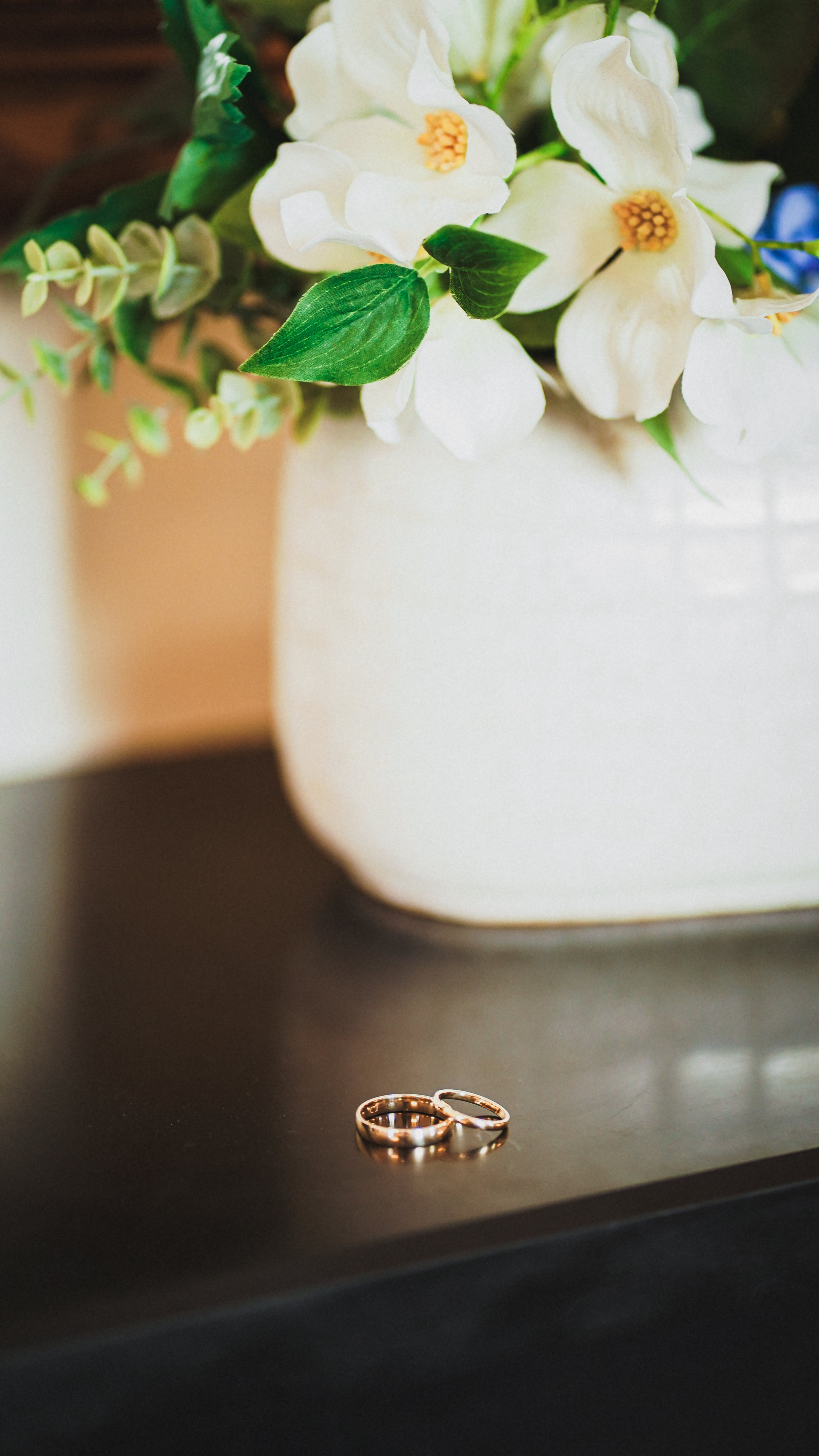 Close-up of two gold wedding rings on a dark surface, with a blurred background of a white vase holding white flowers and green leaves.