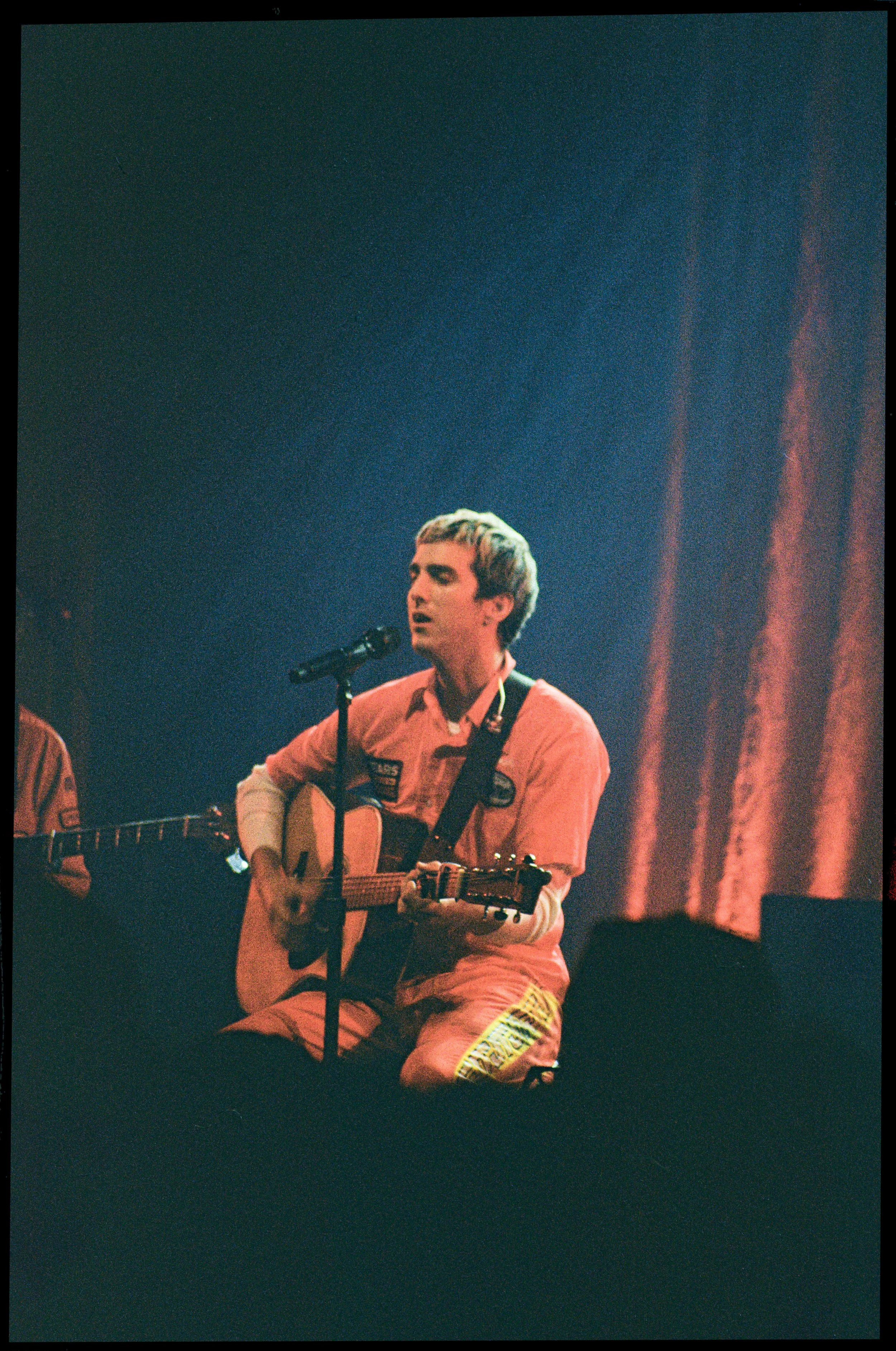 Young man with short hair and light skin, singing into a microphone while playing an acoustic guitar on stage with dim lighting and orange curtains in the background.
