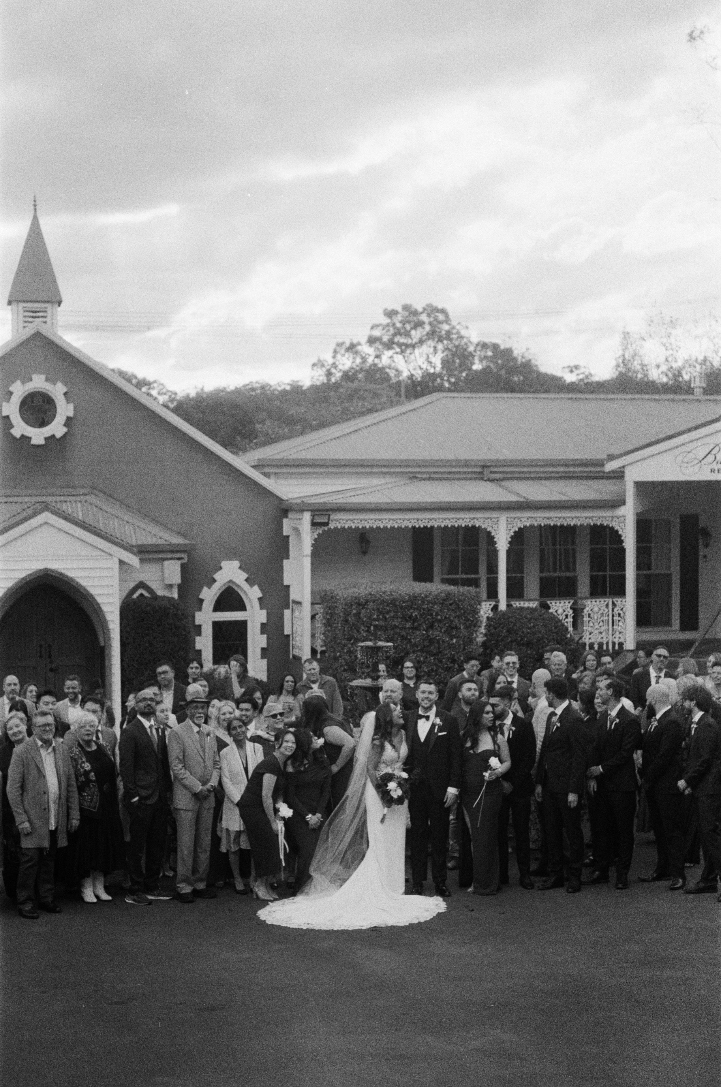 Black and white photo of a wedding celebration outside a church with a large crowd of people, including the bride and groom in the center.