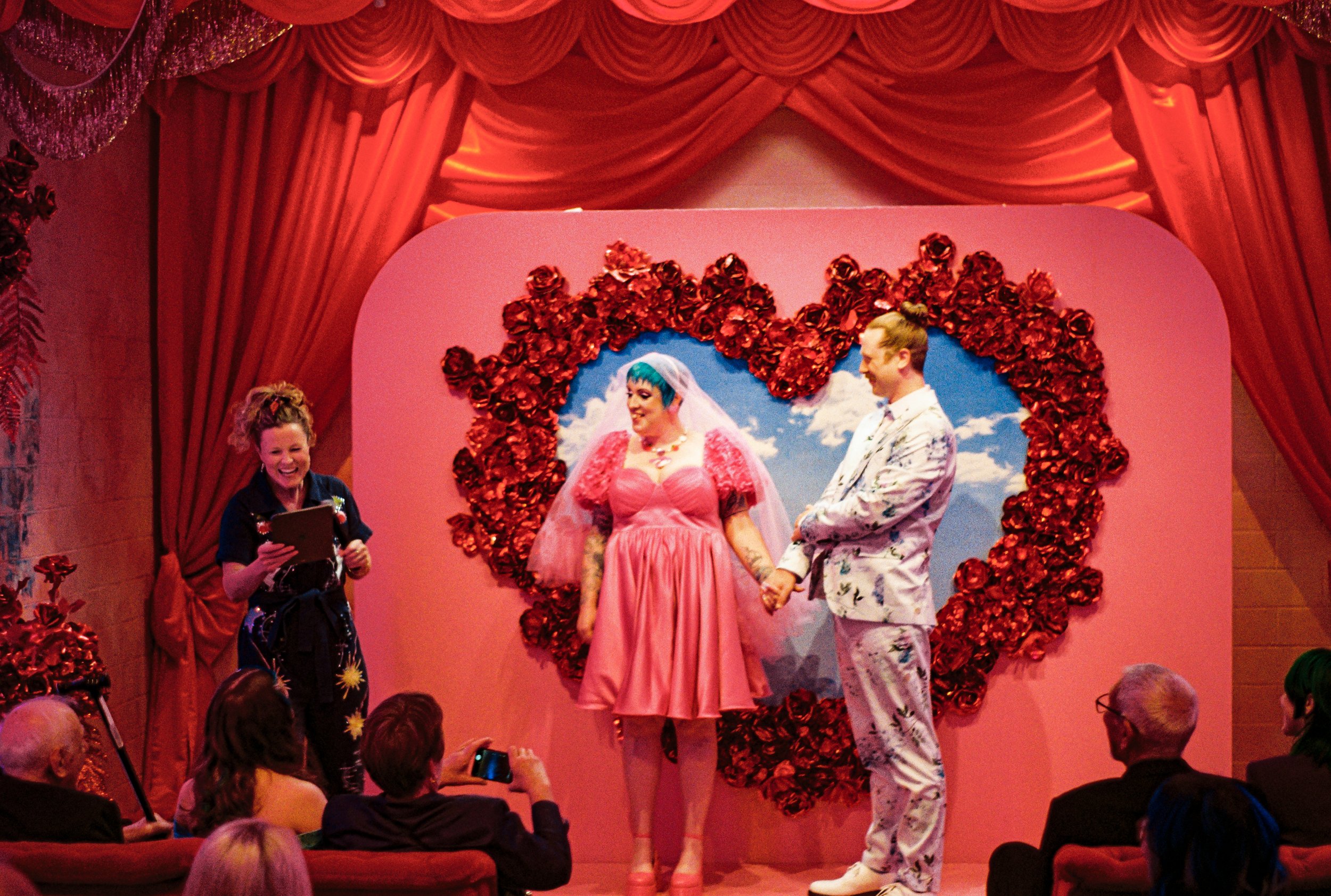 A wedding ceremony taking place on stage with a bride and groom holding hands, surrounded by red roses and a heart-shaped floral arrangement, under red curtains, with guests seated and taking photos.