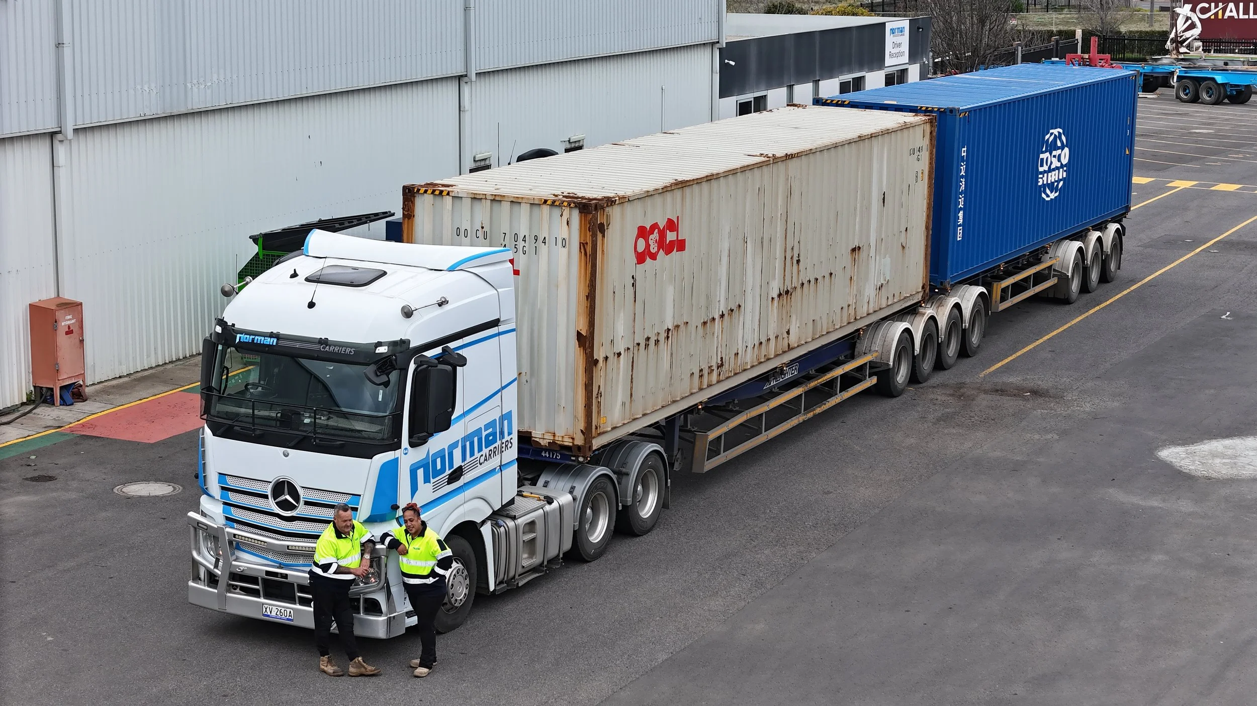 A large white truck with blue lettering that reads 'Norman Carriers' is parked outside a building, with two men in high-visibility jackets standing in front of it, talking. There are two shipping containers on the truck, one beige and rusty, the othe
