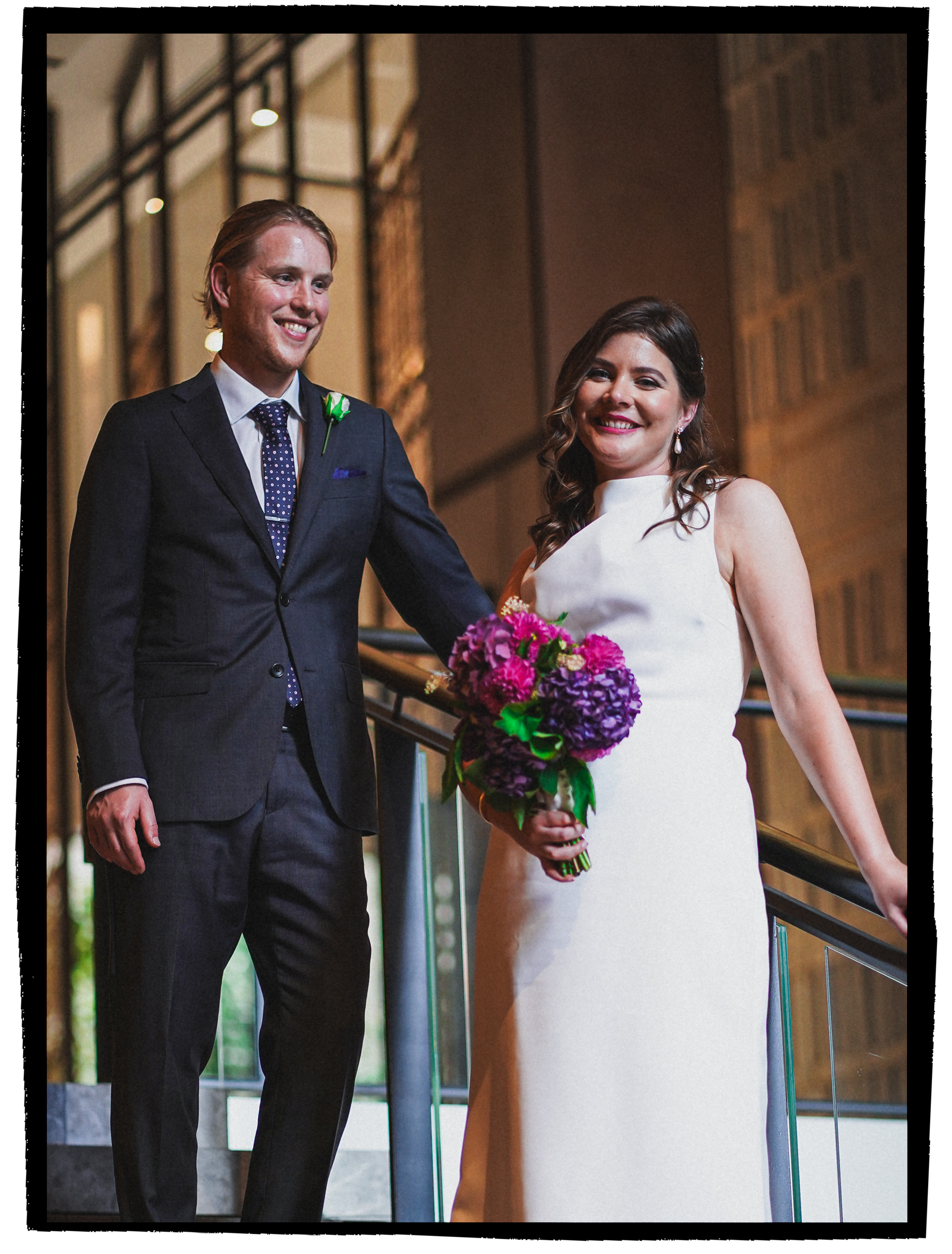 A man and a woman dressed in wedding attire smiling at each other at a wedding reception indoors, with a glass railing and cityscape in the background.