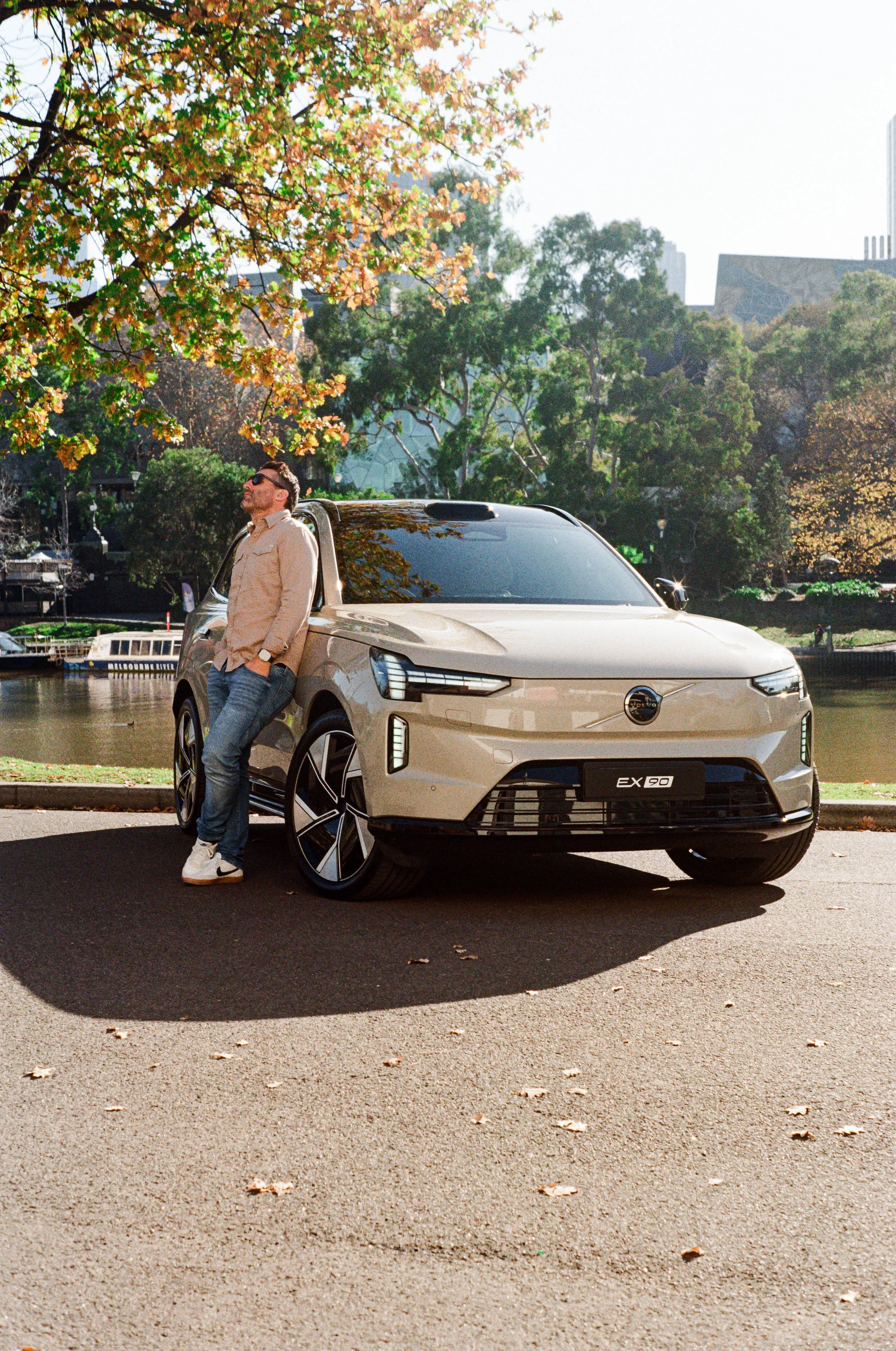 A man leaning against a beige Volvo EX90 SUV parked on a paved street with fallen leaves. The man is wearing sunglasses, a beige jacket, and jeans. There are trees with autumn foliage and a river with boats in the background.