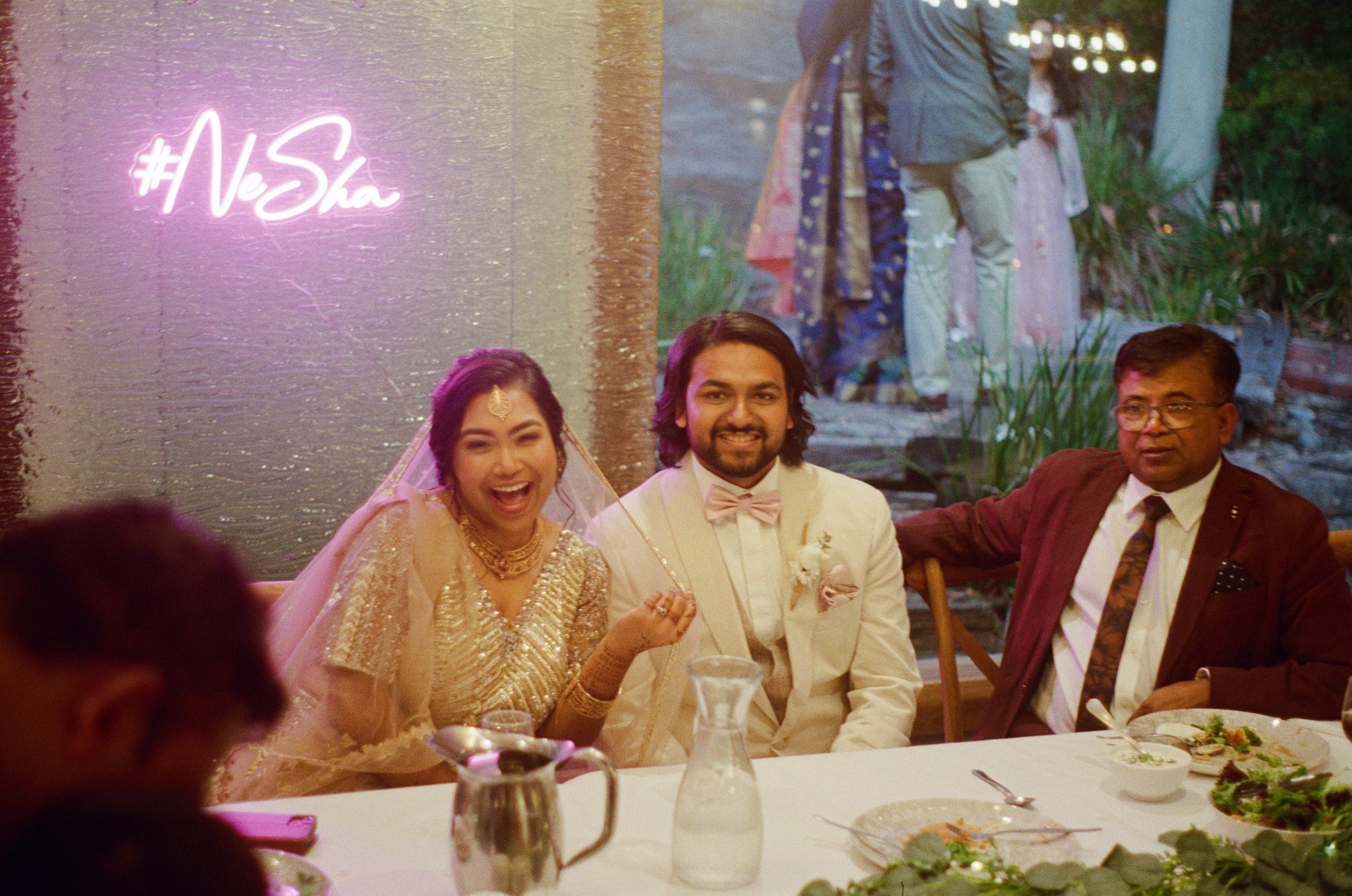 Happy bride and groom sitting at a wedding reception table, smiling, with friends in the background and a neon sign that says '#NeSha' on the wall.