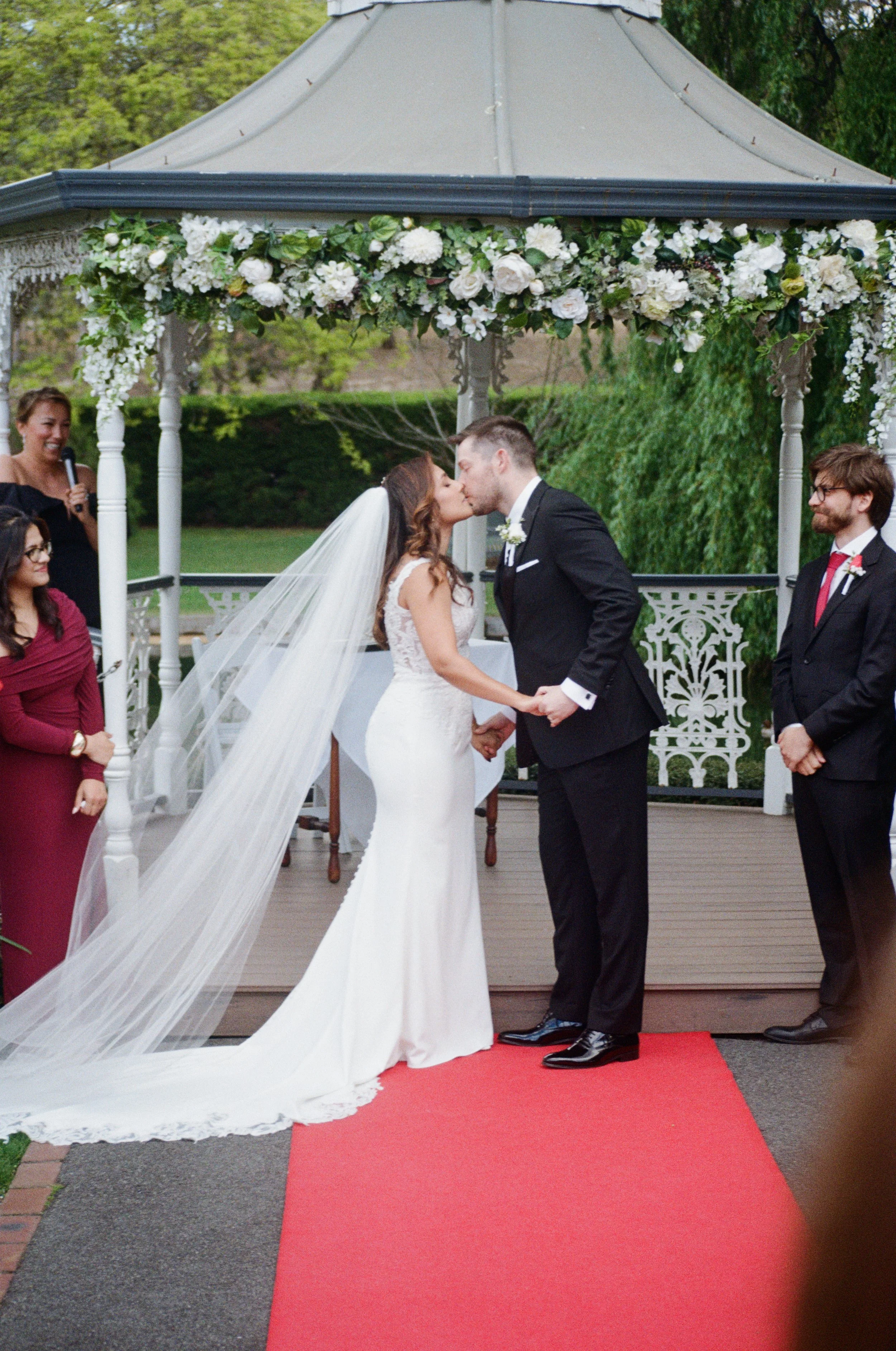 A bride and groom kissing during their wedding ceremony under a decorated gazebo with white flowers. Wedding guests look on, some smiling, on a red carpet outside with trees in the background.