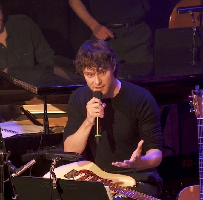 A young man with curly hair is holding a microphone and speaking on stage. He is sitting with a white electric guitar in front of him, surrounded by musical equipment, with a piano and another person in the background.