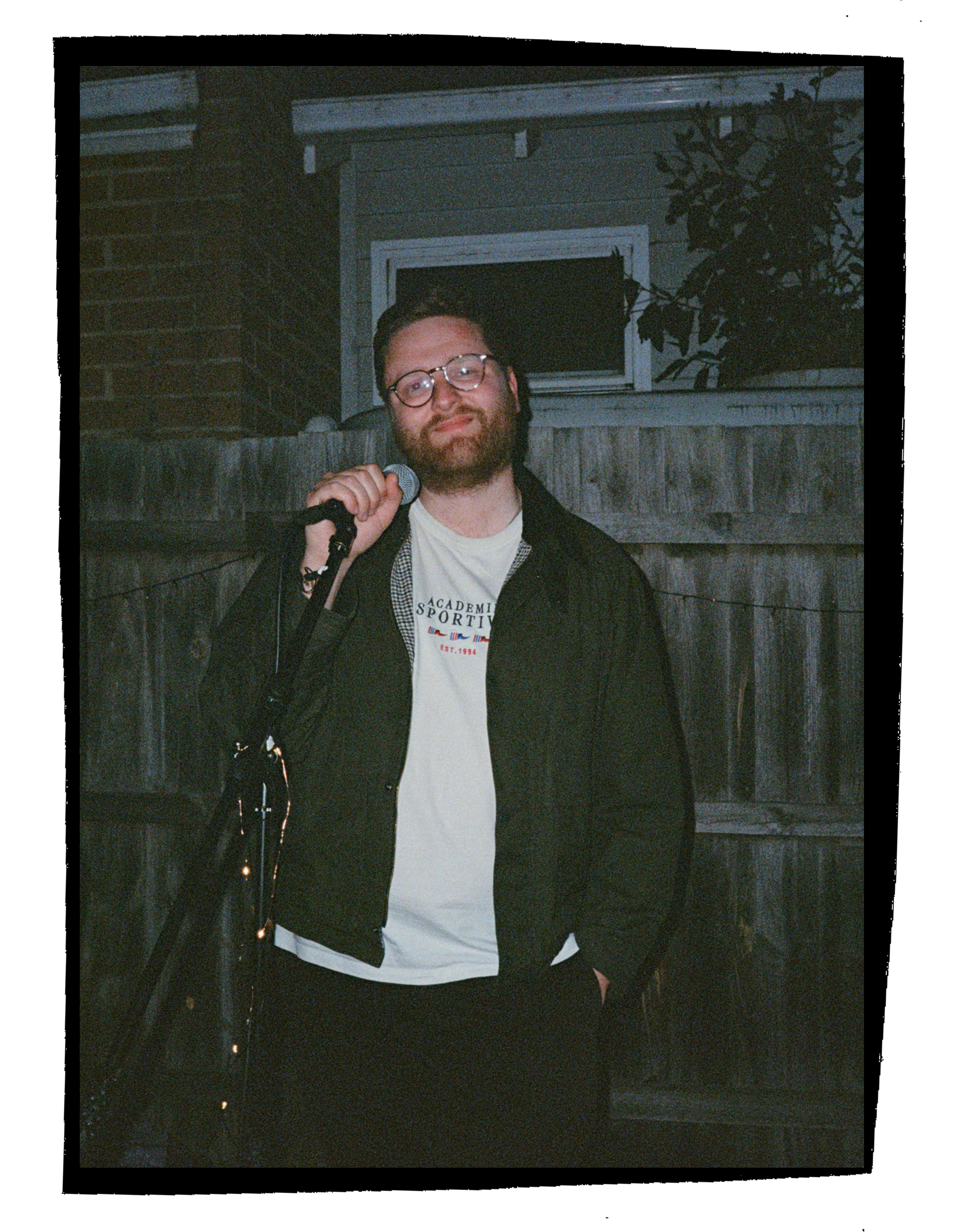 A man with glasses and a beard holding a microphone standing outdoors at night, with a wooden fence and a small window in the background.