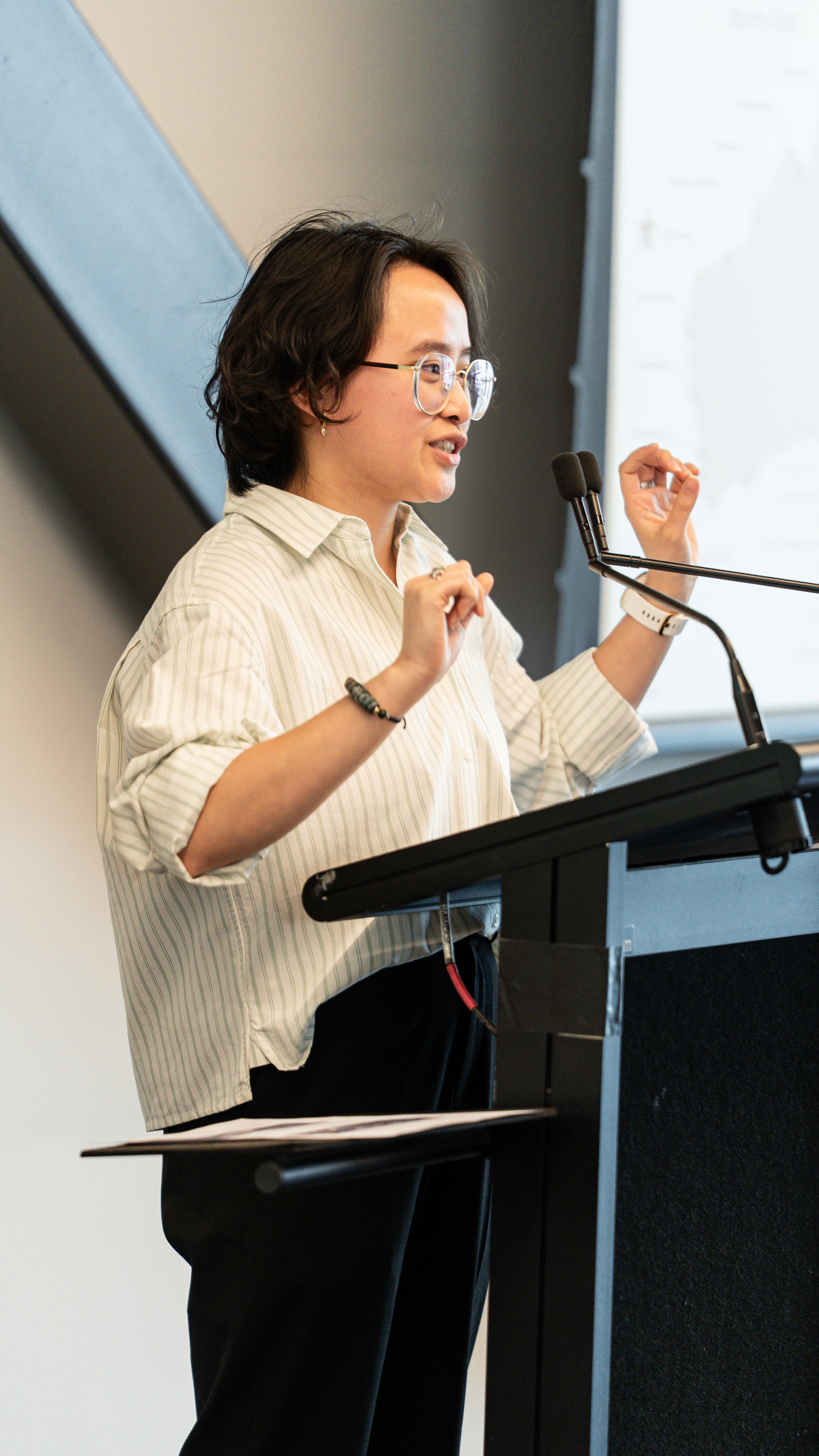 A woman with glasses giving a presentation at a podium with a microphone, wearing a striped shirt and black pants.