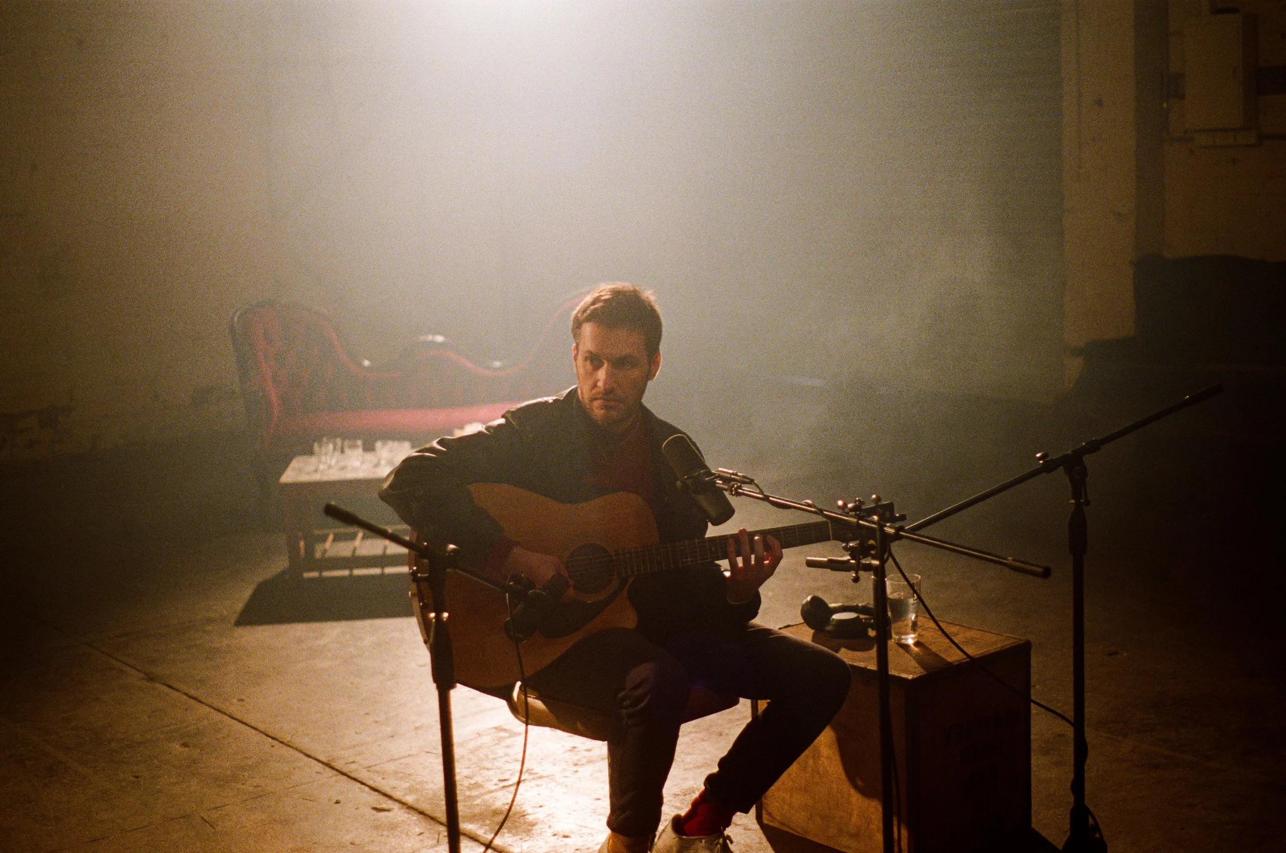 A man playing an acoustic guitar and singing into a microphone on a dimly lit stage with a vintage-style chair and piano in the background.