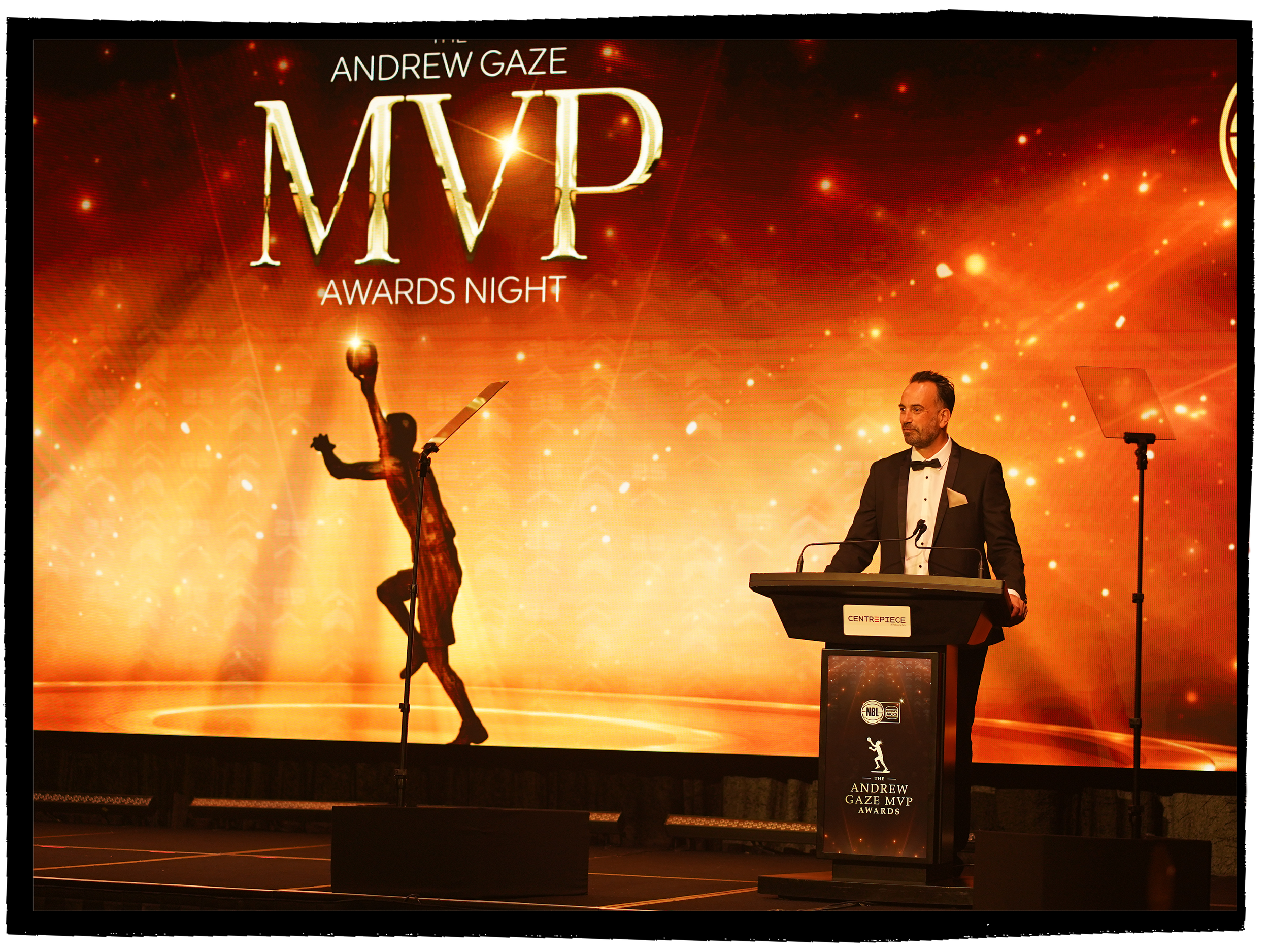 Man in tuxedo speaking at podium during awards ceremony with a large screen behind displaying event information, including the name 'Andrew Gaze MVP MVP Awards Night' and a silhouette of a basketball player against an orange and red background with s