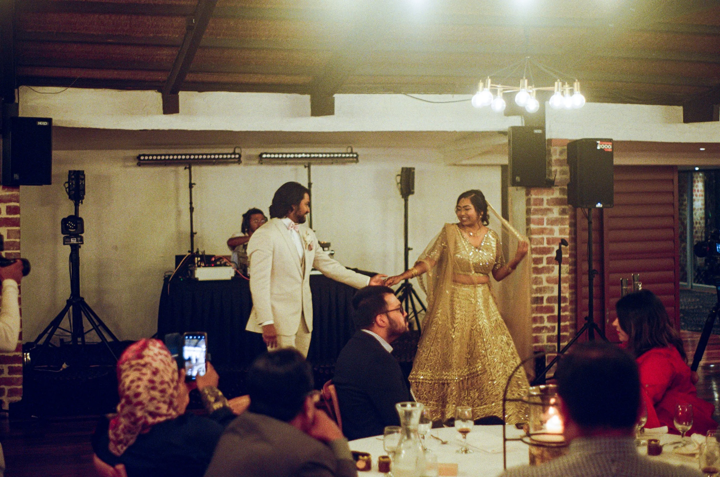 A man and woman dressed in traditional South Asian wedding attire are holding hands on the dance floor at a wedding reception. The man is in a cream-colored suit, and the woman is in a golden lehenga. They are smiling and looking at each other, with 