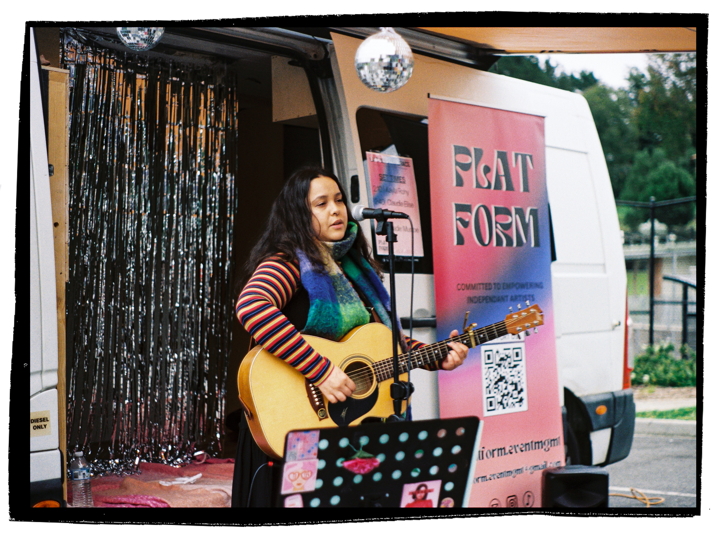 A woman playing guitar and singing into a microphone outdoors during daytime, with informational banners and decorations, including a disco ball hanging above.