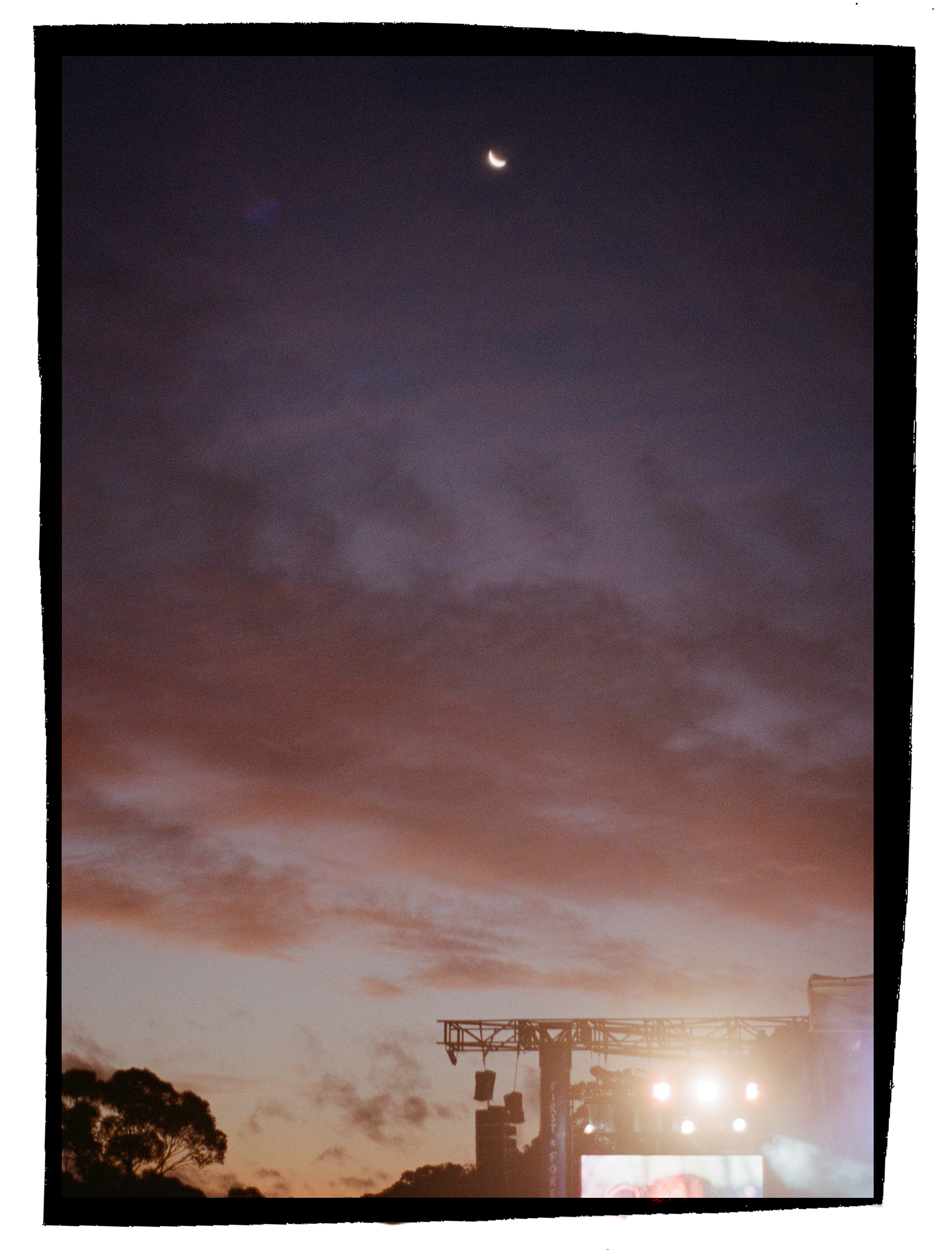 Photo of a sunset sky with clouds and a crescent moon above a stage setup with lighting equipment, trees, and a billboard at the bottom.