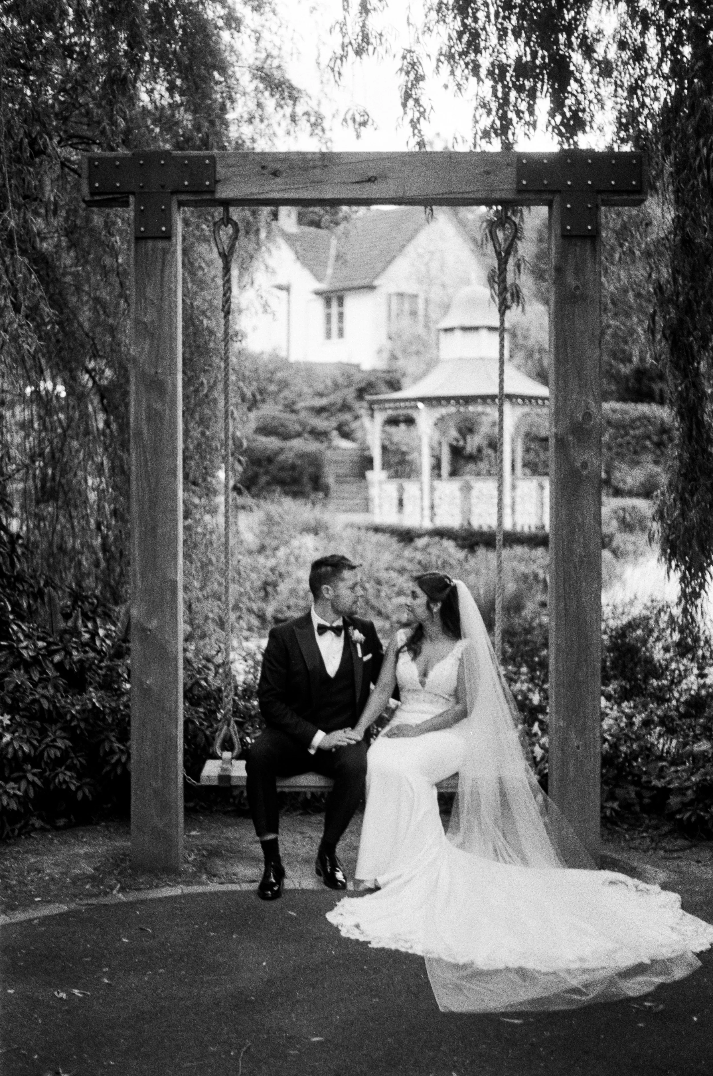 A bride and groom holding hands and kissing under a wooden archway with a swing, in an outdoor garden setting.