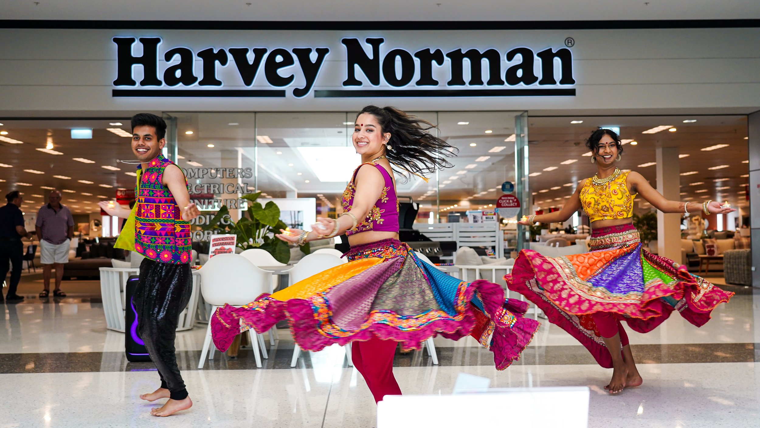 Colorful traditional Indian dance performance with three dancers in vibrant attire inside a shopping mall entrance with the Harvey Norman sign.