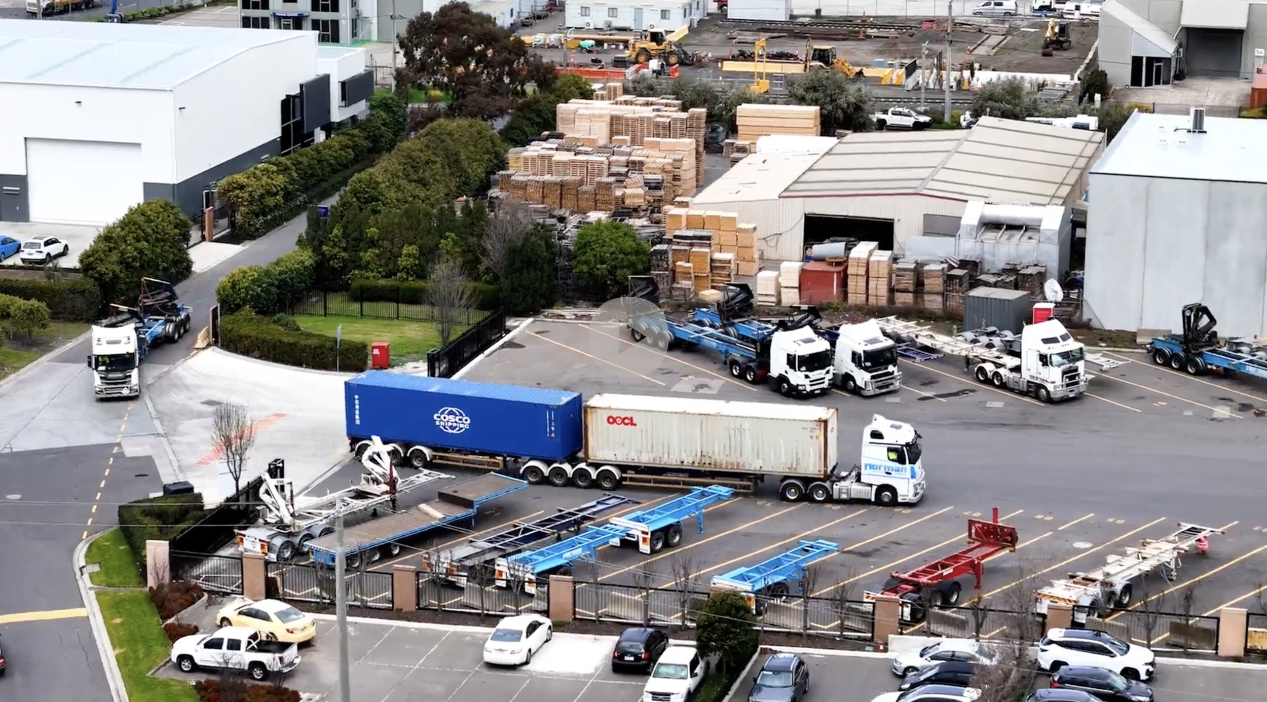 View of a parking lot and industrial area with trucks, trees, and buildings. Several trucks are parked or moving, with one carrying blue construction equipment. There are stacks of building materials, construction equipment, and a fenced area with sm