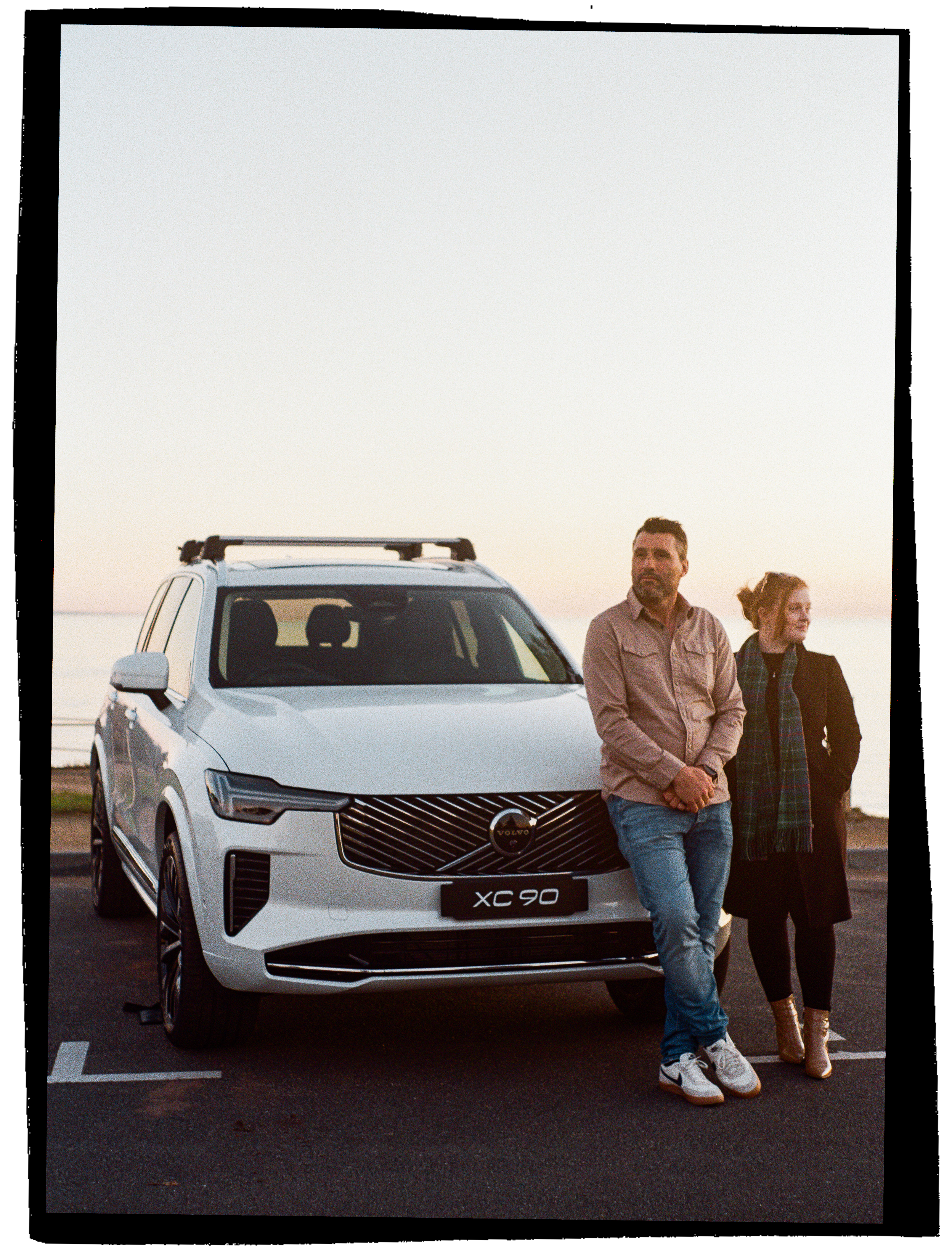 A man and a woman standing in front of a white Volvo XC90 SUV during sunset at the beach.