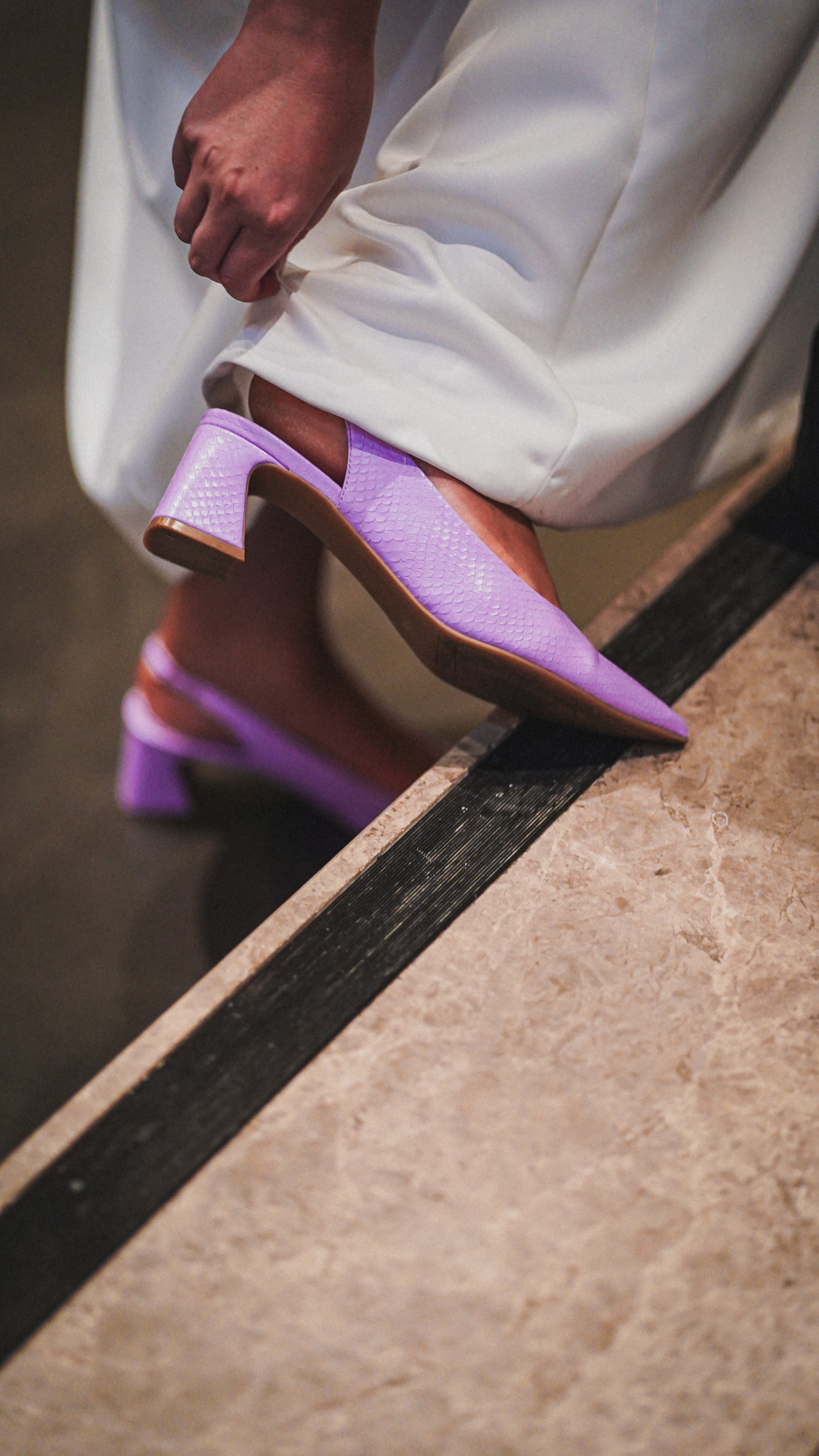 A person wearing white pants putting on purple high-heeled shoes with a snakeskin pattern.