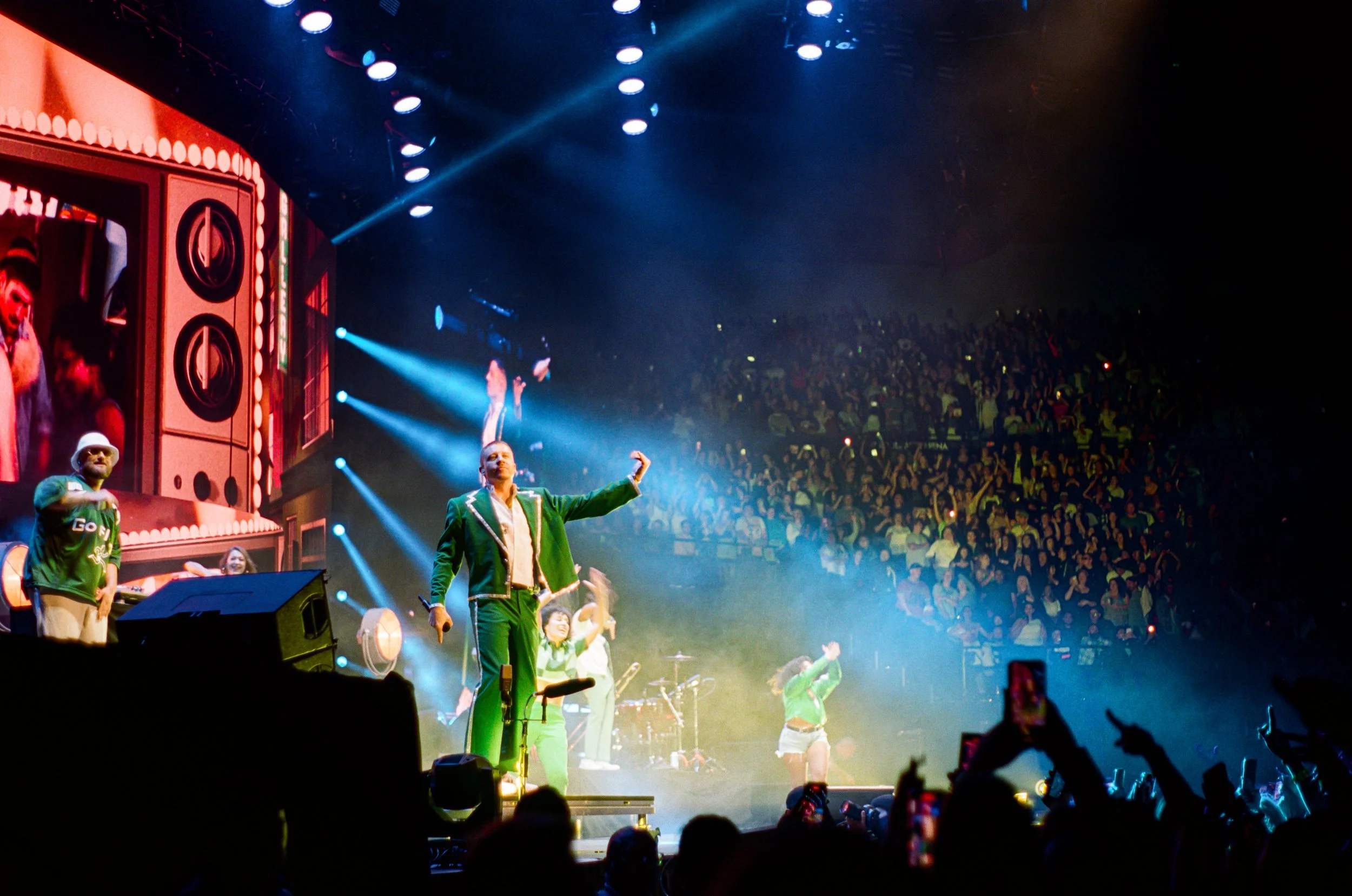 A lively stage performance with performers in green outfits, some with microphones, in front of a large cheering audience. The stage is lit with bright blue and white lights, and a big screen shows a close-up of some performers.