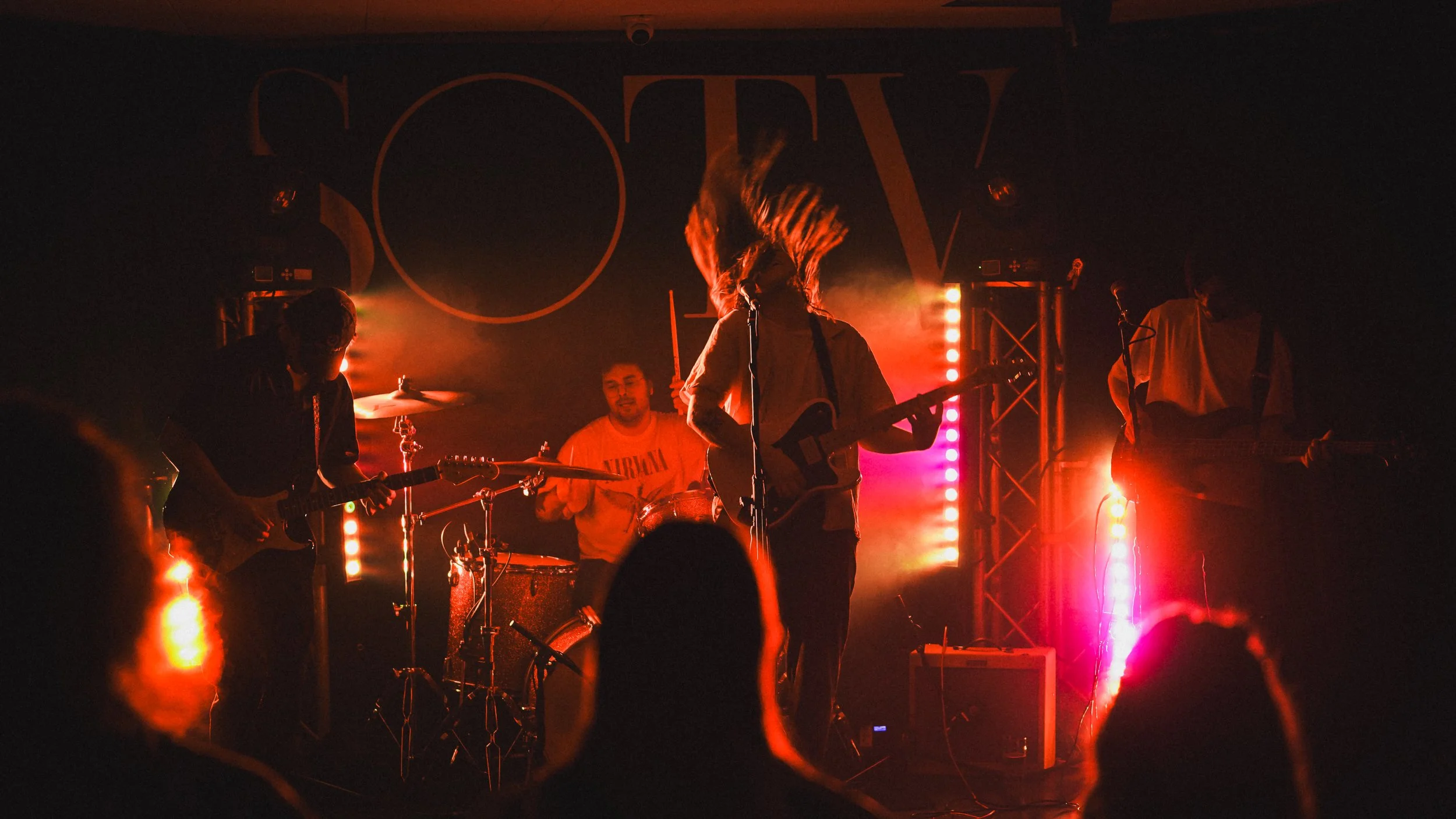 A live band performance on stage, with band members playing guitars and drums under colorful stage lighting including red, purple, and yellow hues.