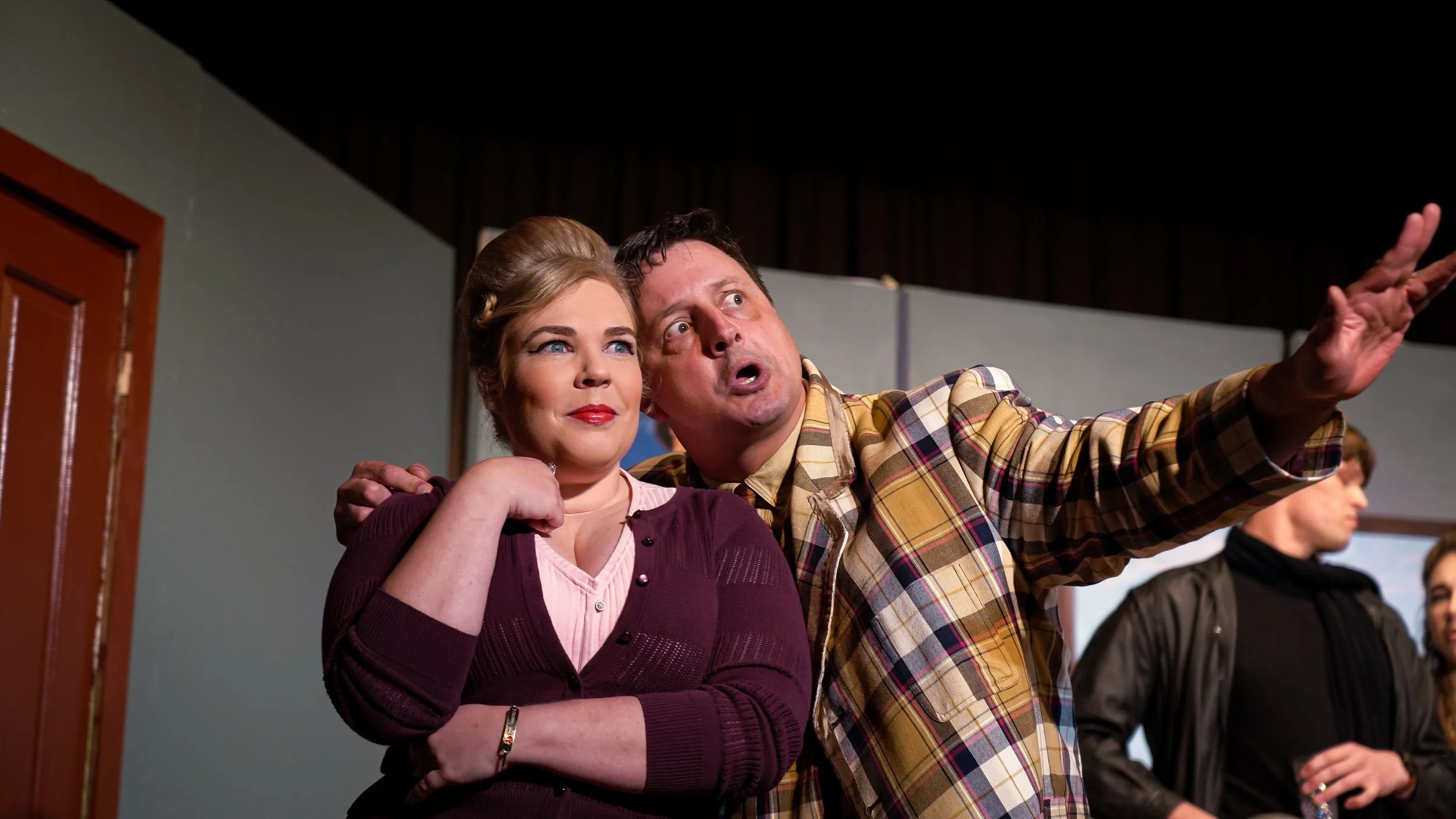 Two actors on stage, one woman and one man, appear to be acting in a theatrical performance. The woman has blonde hair styled in vintage waves and wears a maroon cardigan over a pink blouse. The man, with dark hair, is wearing a plaid jacket and appe