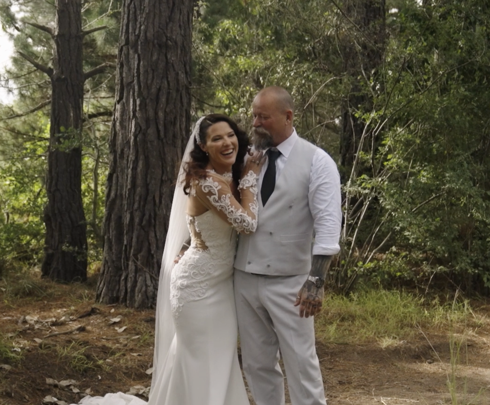 A bride and groom holding each other and smiling in a forest setting on their wedding day, with tall trees and lush greenery in the background.