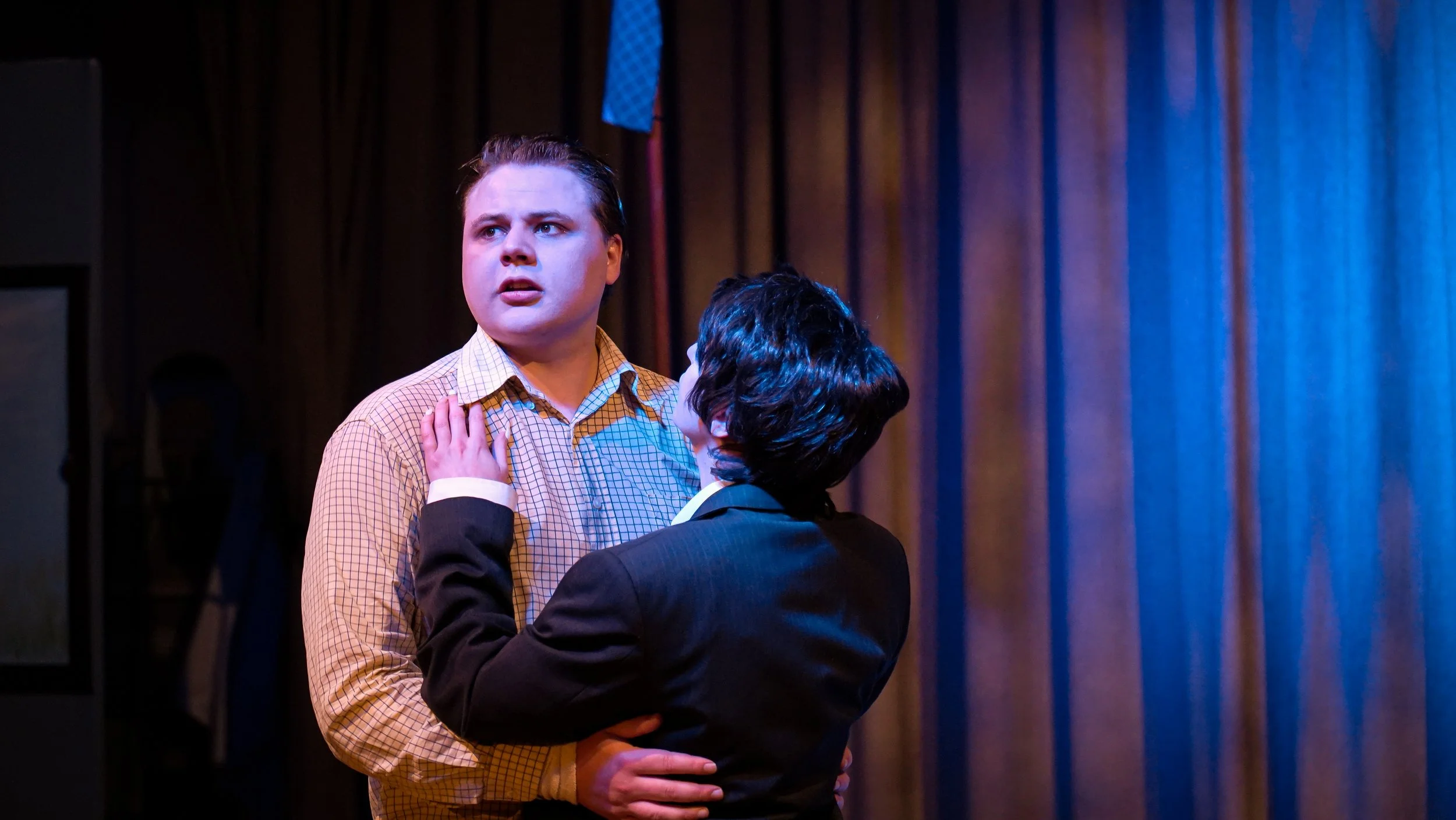 Two men are on a stage, one looking surprised or worried, and the other holding his waist with both hands. The man facing the camera has light-colored hair and is wearing a checkered shirt, while the other has dark hair and is wearing a dark suit. Th
