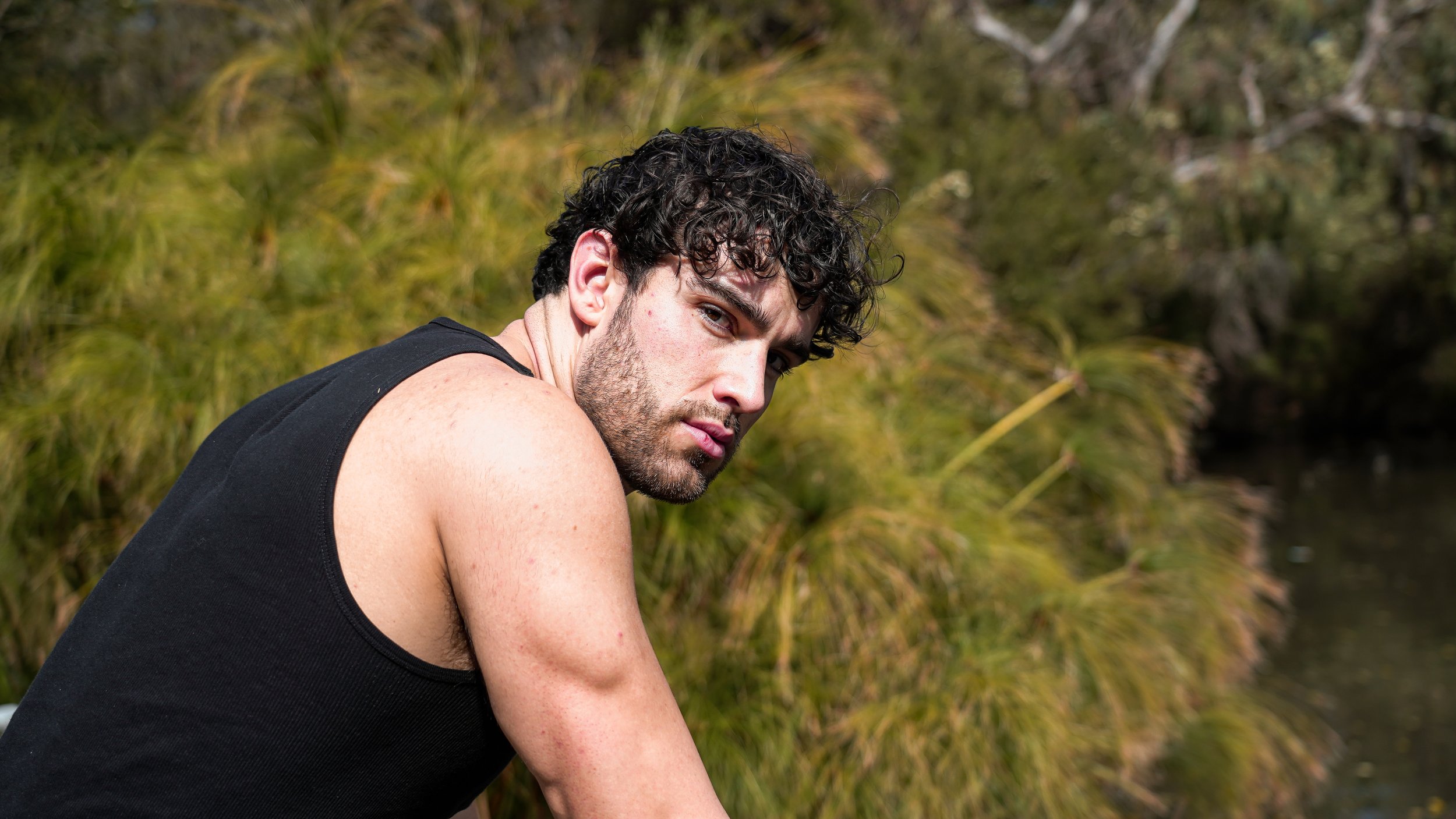 A young man with curly dark hair and a black sleeveless shirt looking over his shoulder outdoors.