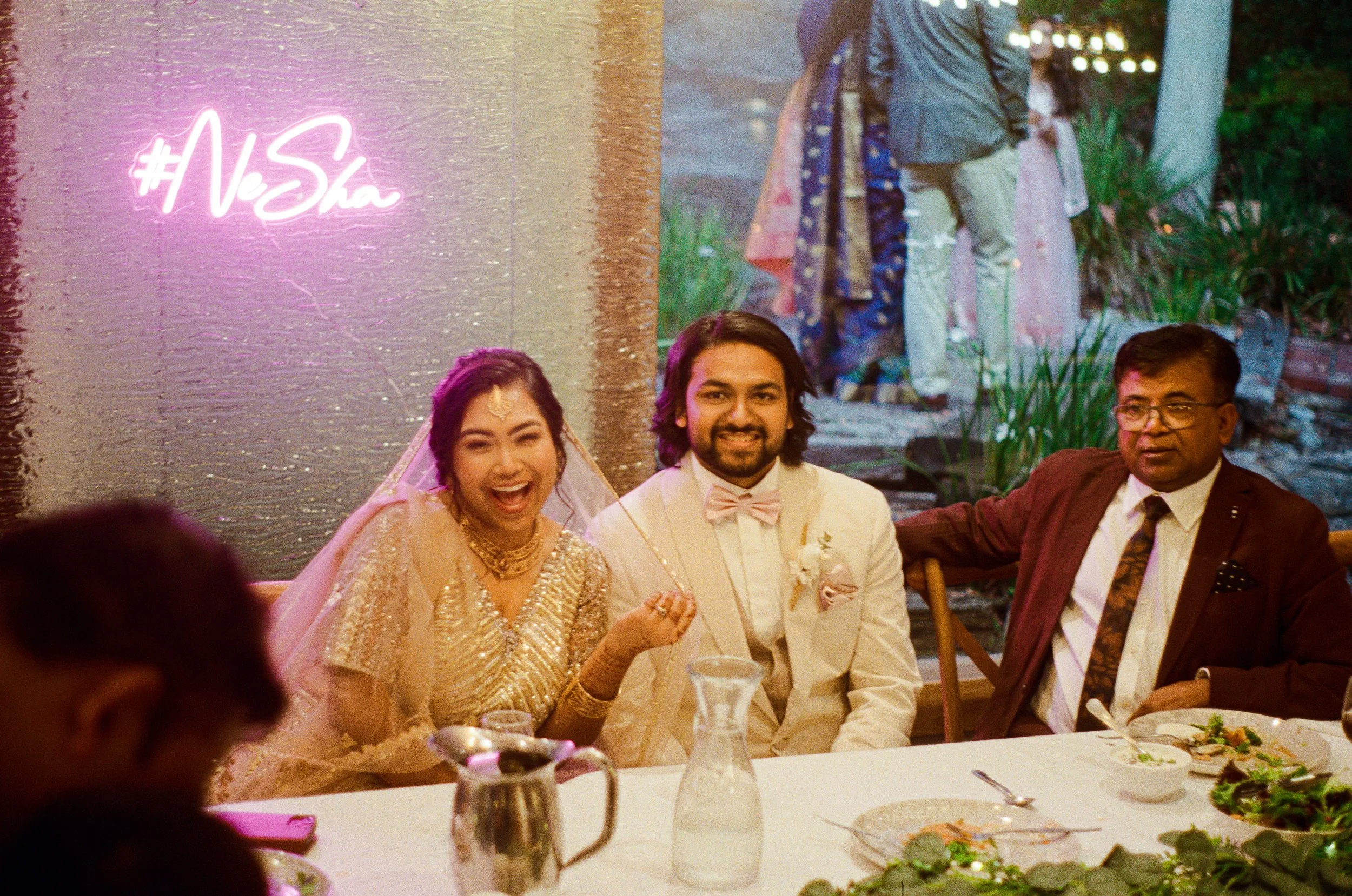 People dressed in formal attire at a wedding reception, sitting at a table with plates of salad and wine glasses, with a neon sign that reads '#VeSha' on the wall behind them.