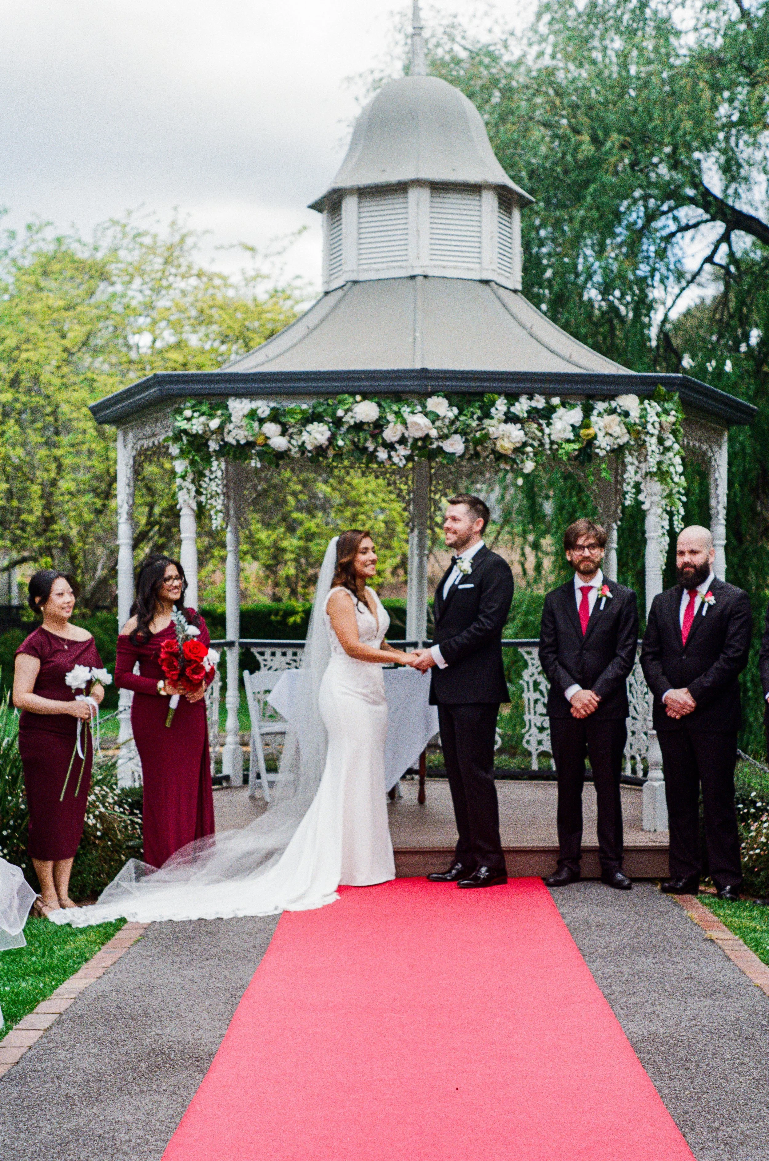 A wedding ceremony taking place outdoors under a decorated gazebo, with a bride and groom holding hands and smiling at each other, surrounded by attendants, on a red carpet.