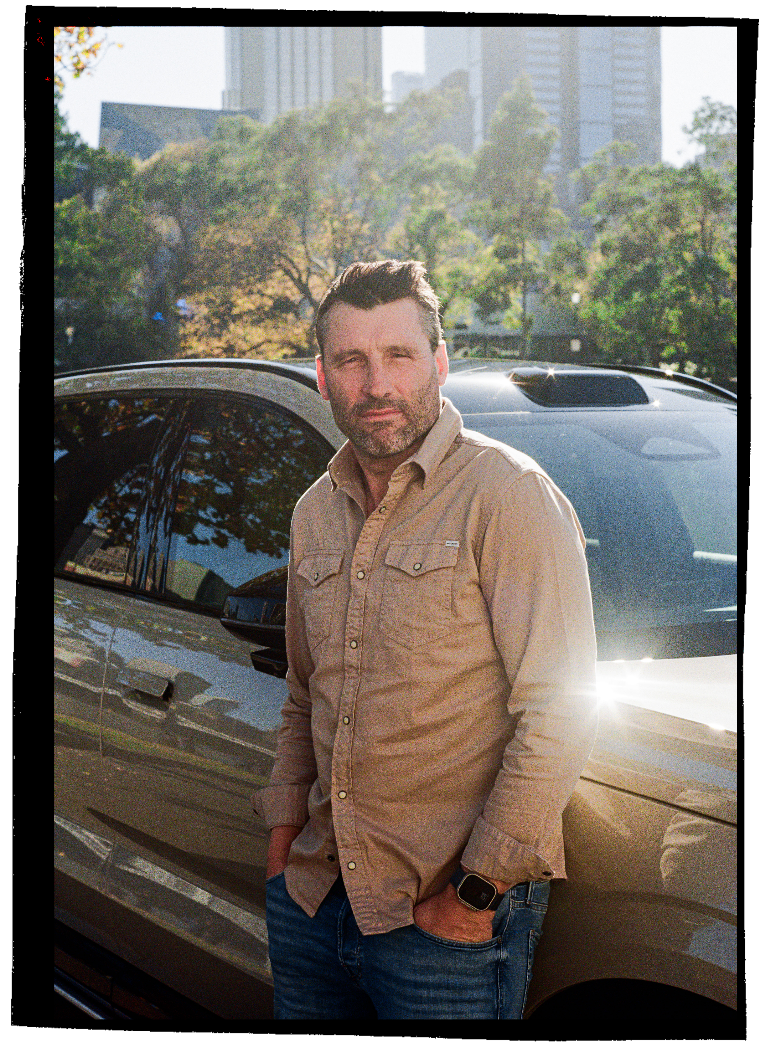 A man with dark hair and a beard standing next to a parked car, with a city skyline and trees in the background, as the sun sets.