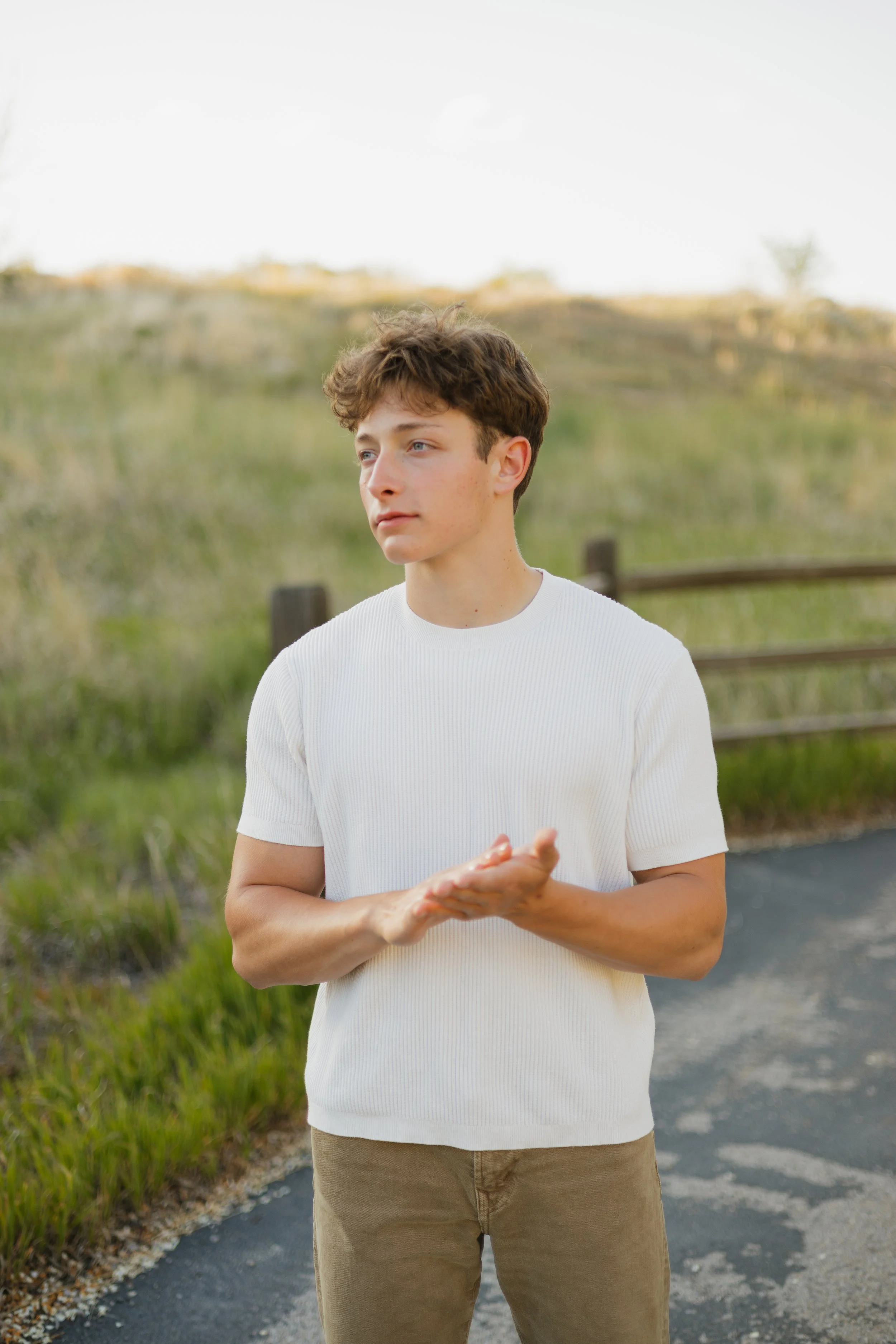 A young man with curly brown hair and fair skin standing outdoors on a paved path, wearing a white short-sleeved shirt and tan pants, looking to the left with a thoughtful expression, with a rural landscape and wooden fence in the background under an overcast sky.