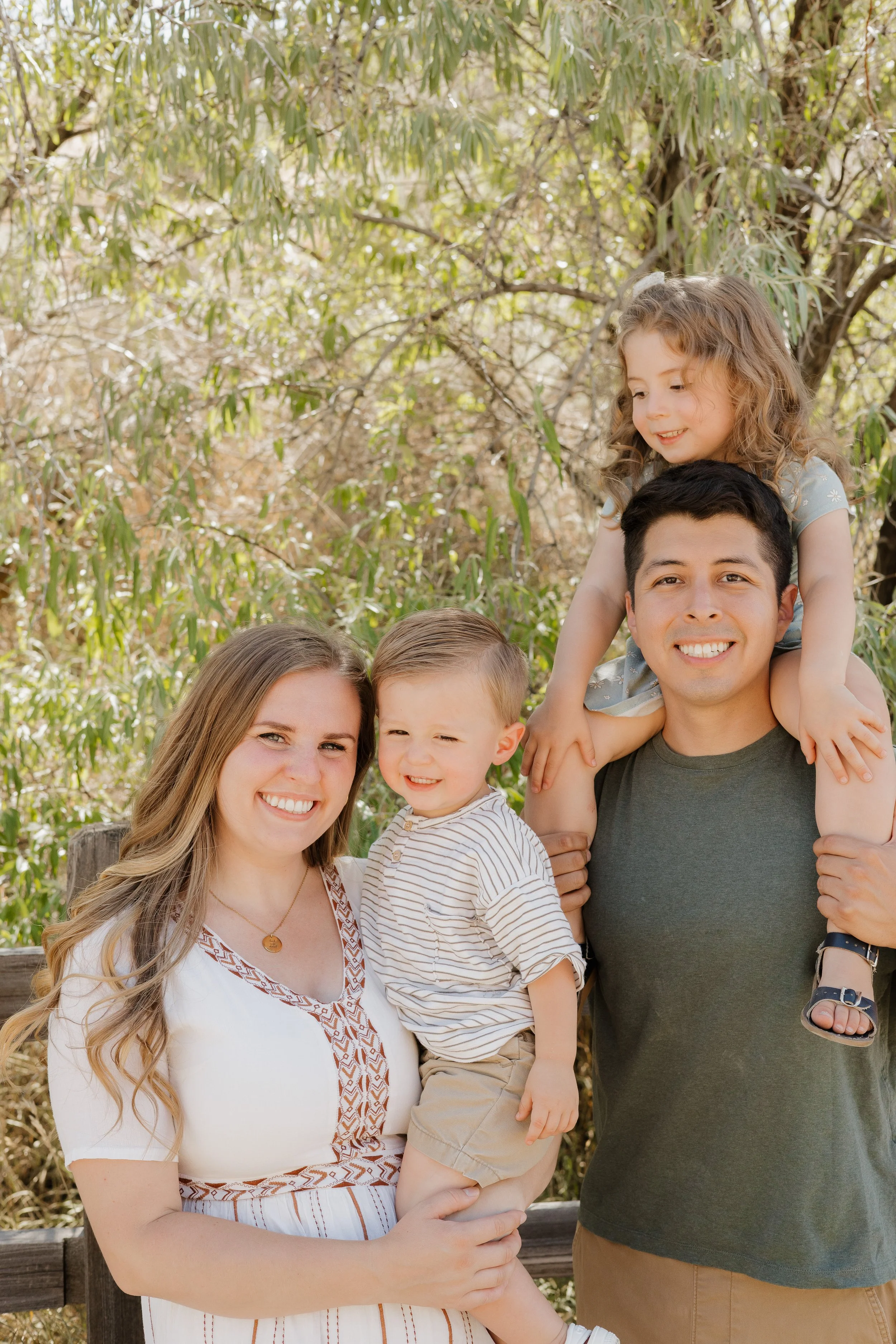 A family of five enjoying fall foliage on a wooded trail, with yellow leaves scattered on the ground. The adults and children are smiling and interacting together.