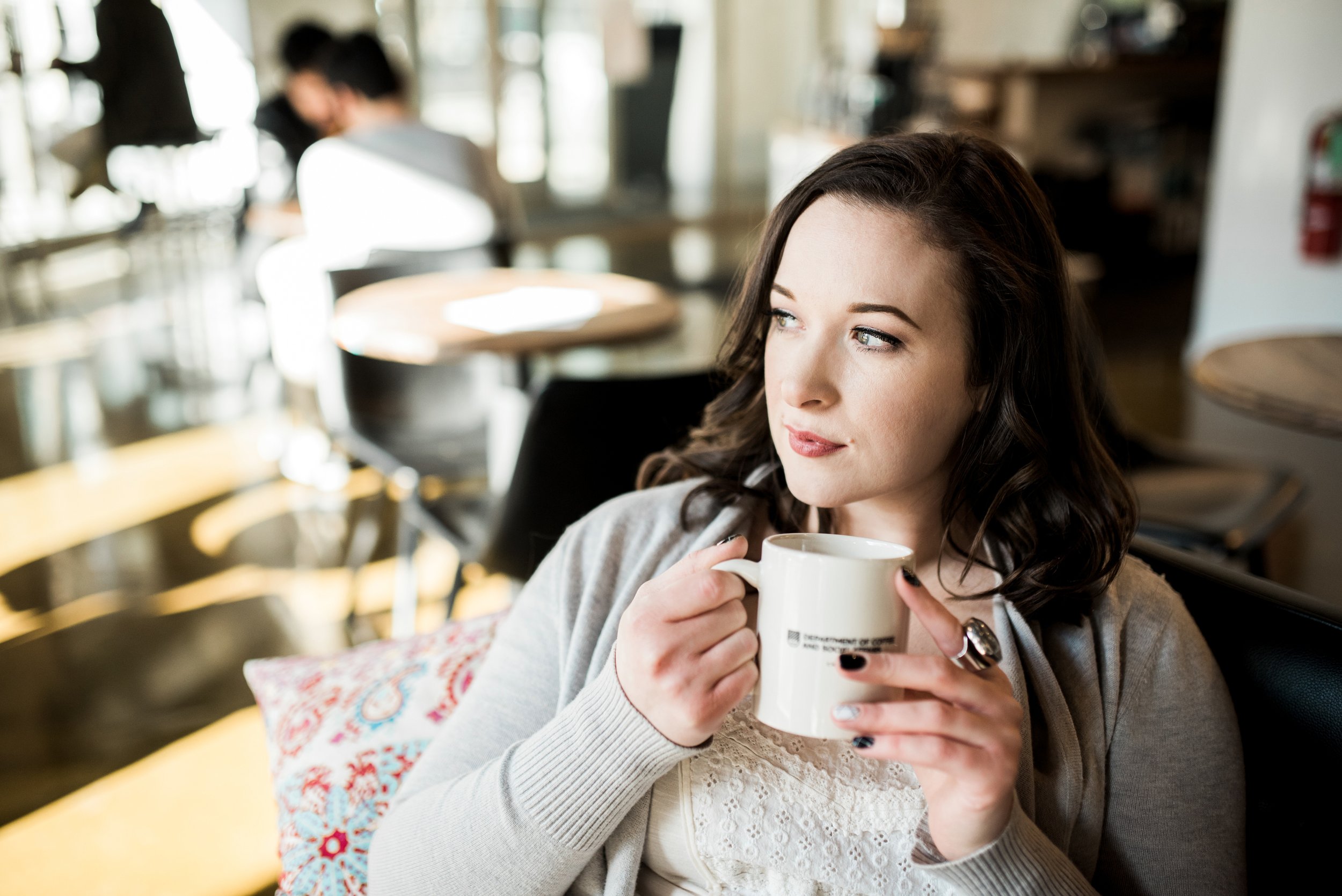 A woman with dark wavy hair holding a white mug, sitting in a cafe with sunlight streaming in, while blurred figures are in the background.