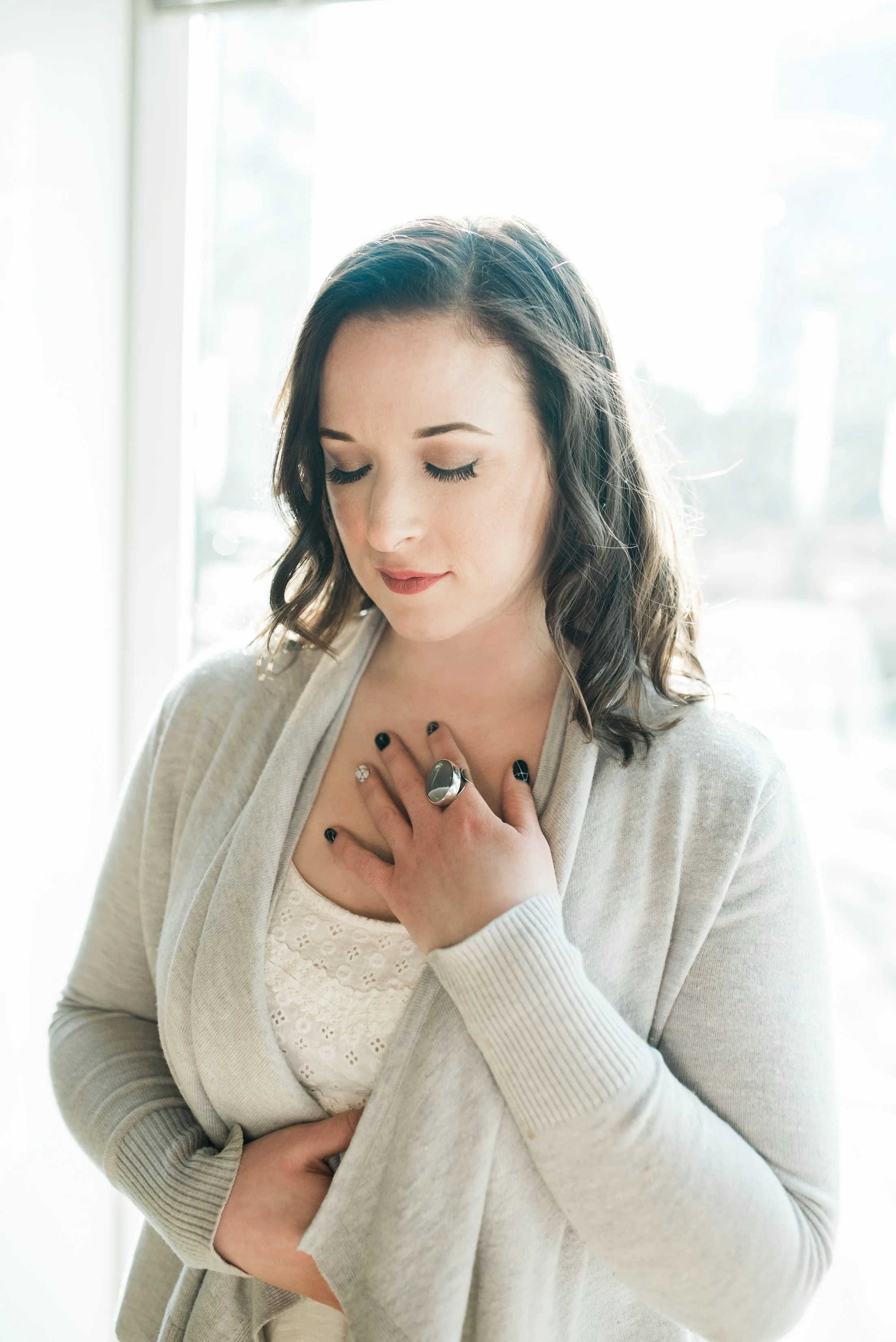 A woman with dark wavy hair, wearing a light beige cardigan and white top, has her eyes closed and is touching her chest with her right hand, which has black nail polish and a large silver ring. She appears to be in a moment of reflection or prayer, standing in front of a bright window.