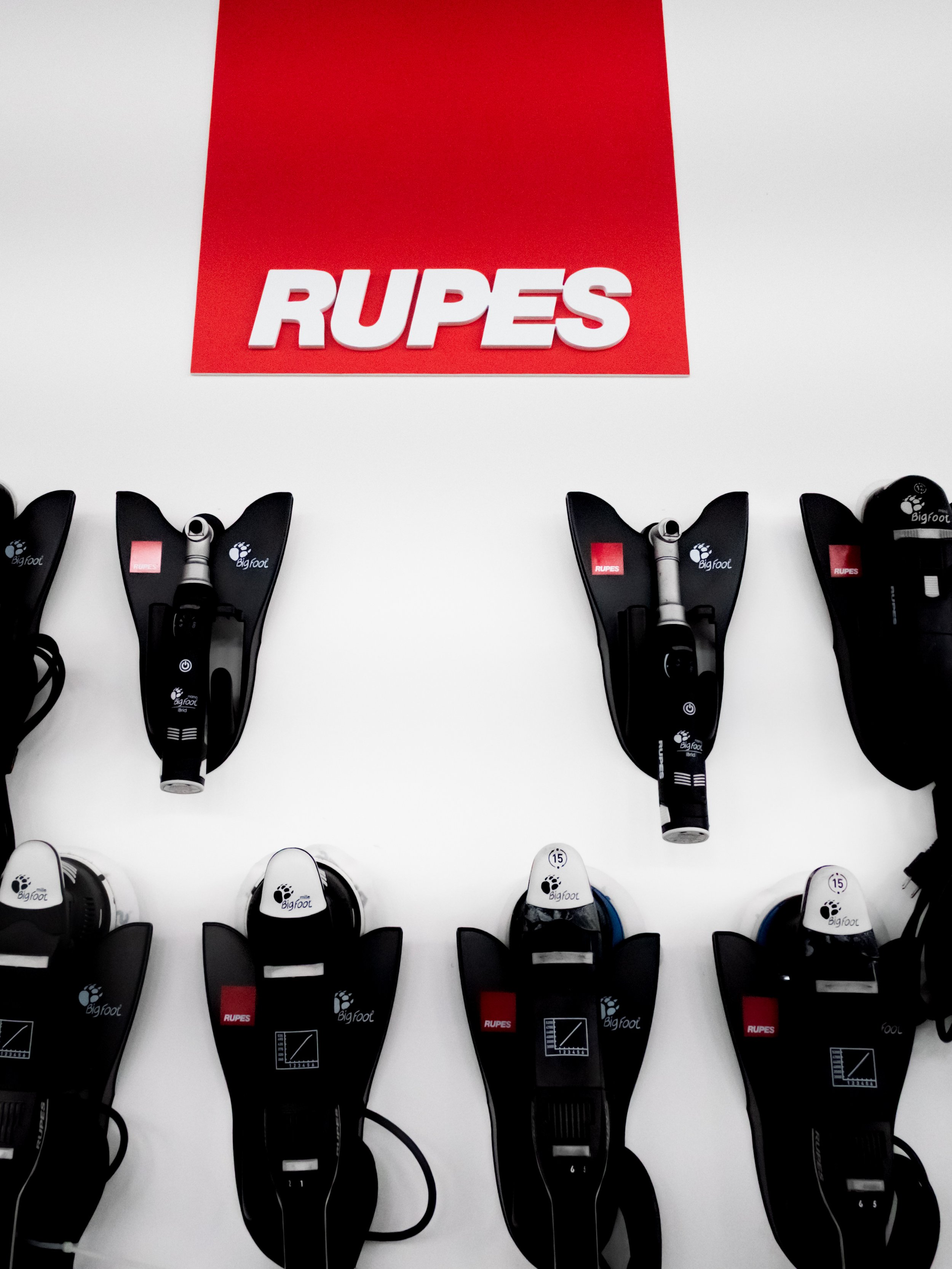 Rows of car detailing hand held polishers used to paint correct vehicles are mounted on a white wall, with a red banner above displaying the word 'RUPES' in white letters.
