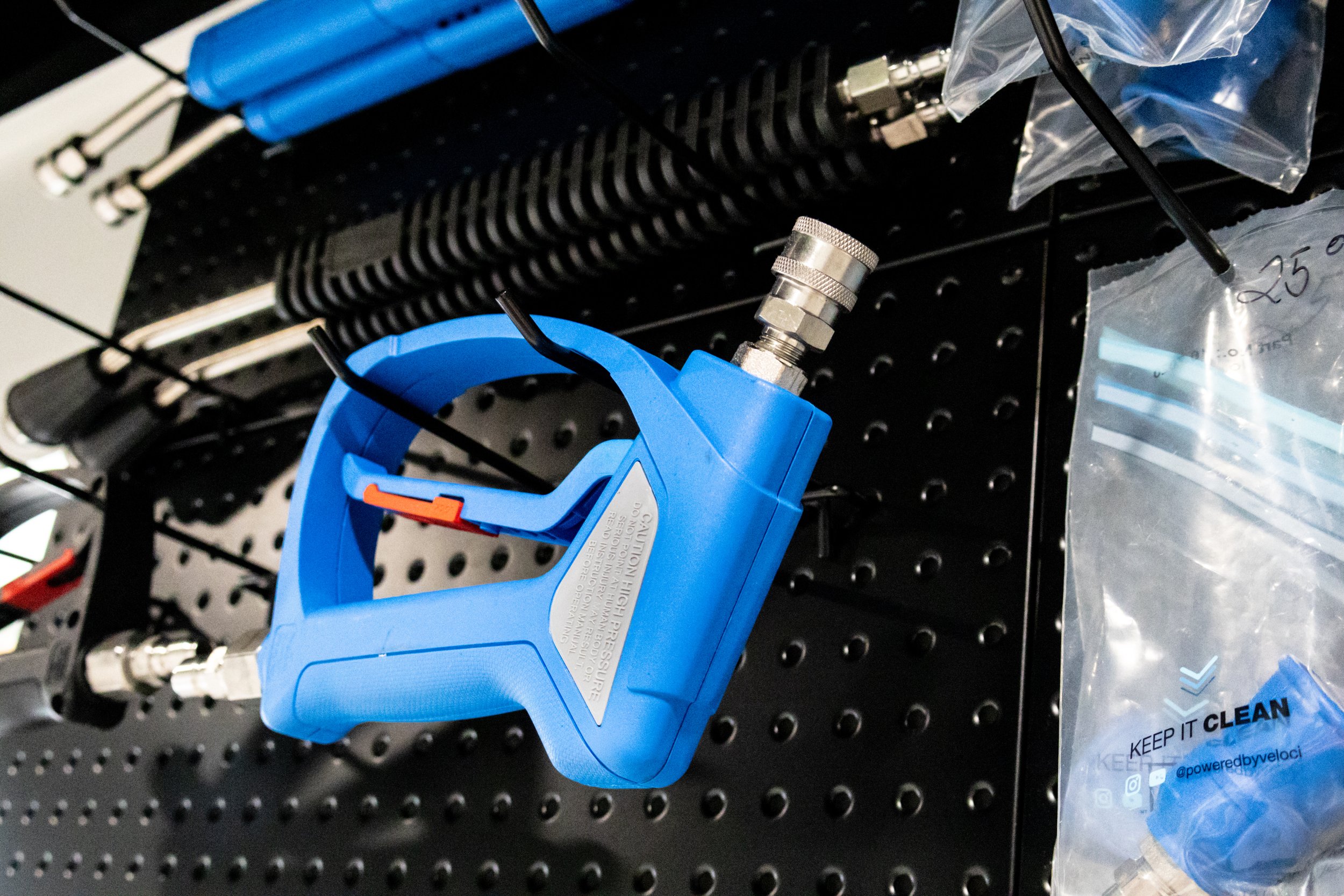 A blue MTM Hydro spray gun attachment used to connect to a foam cannon to wash a car, they are hanging on a black pegboard, with other car detailing hoses and tools and Nozzles nearby.