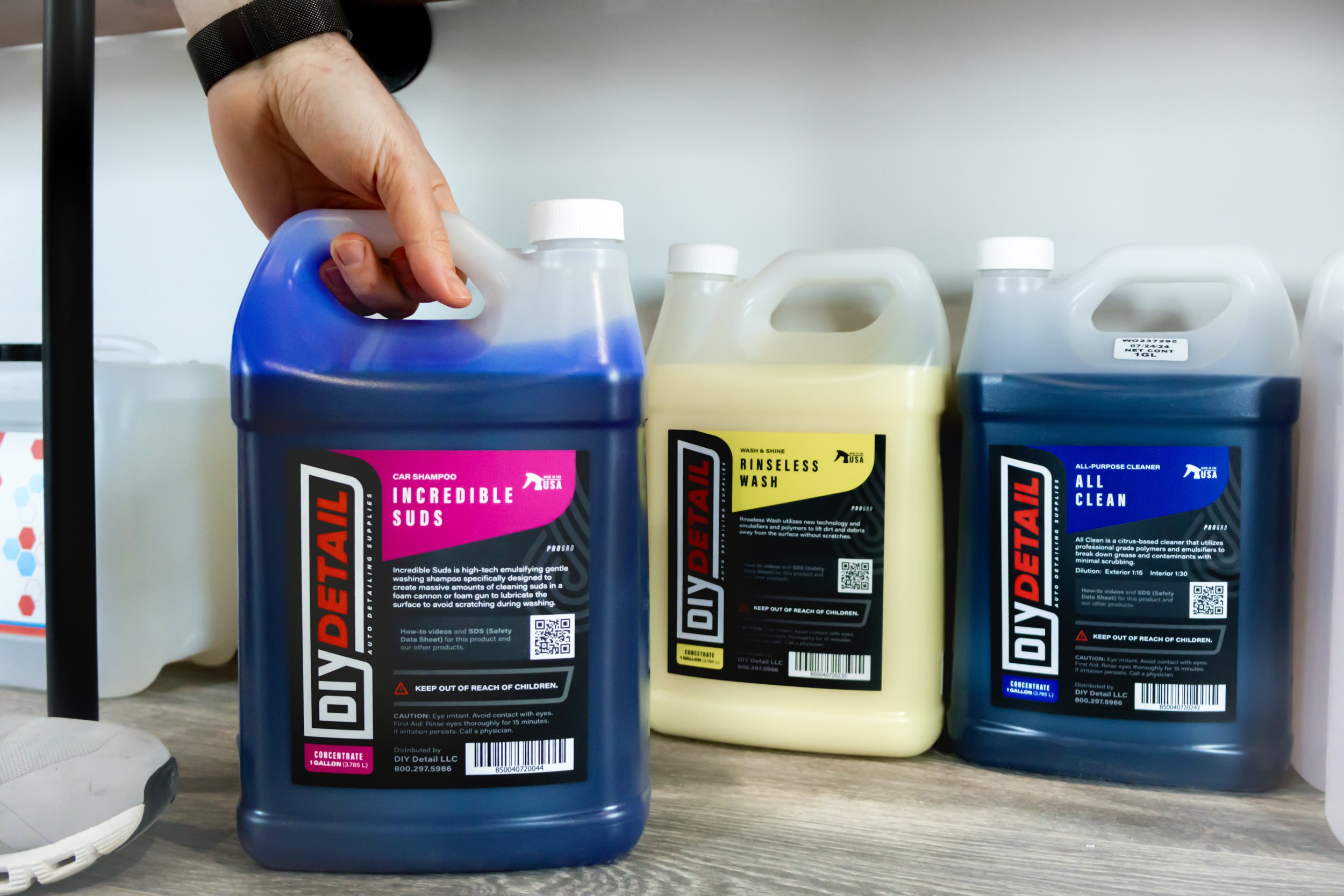 Hand reaching for a gallon of incredible suds, a DIY Detailing car shampoo alongside a gallon of rinsless wash and all clean. Which are detailing products and chemicals used as a multipurpose detailing solution.