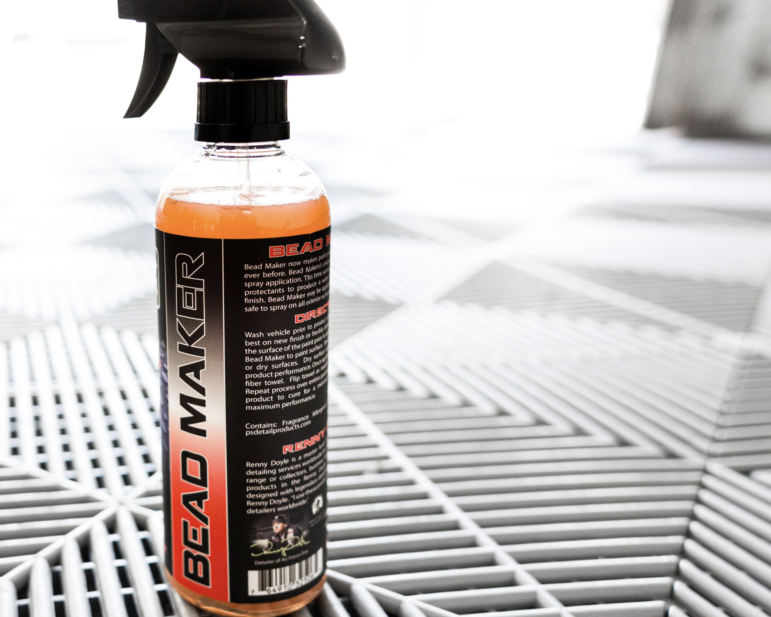 A spray bottle labeled 'BEAD MAKER' containing a detailing solution and sealant used to protect paint on a car. The 16oz bottle with black sprayer is placed on a black and grey racedeck flooring system.