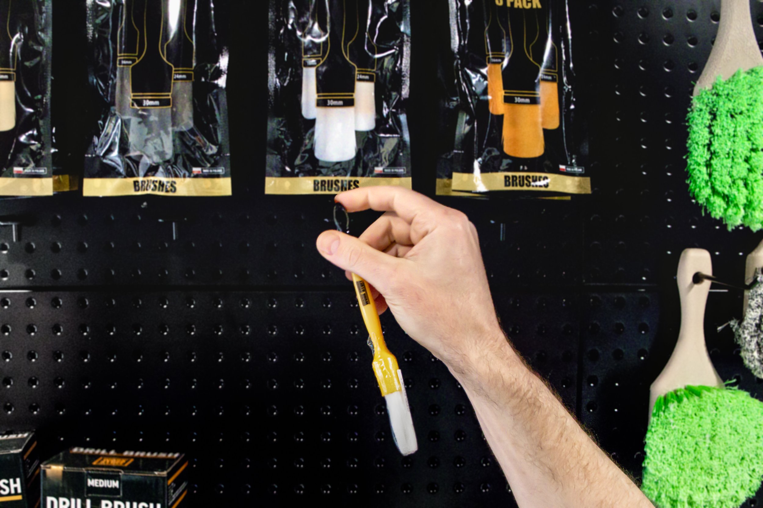 A person holding a Work Stuff Brush with a black handle and white bristles, standing in front of a display of Work Stuff Brushes in black and yellow packaging sold as individual or packs. SM Arnold green tire brushes are on a black pegboard wall.