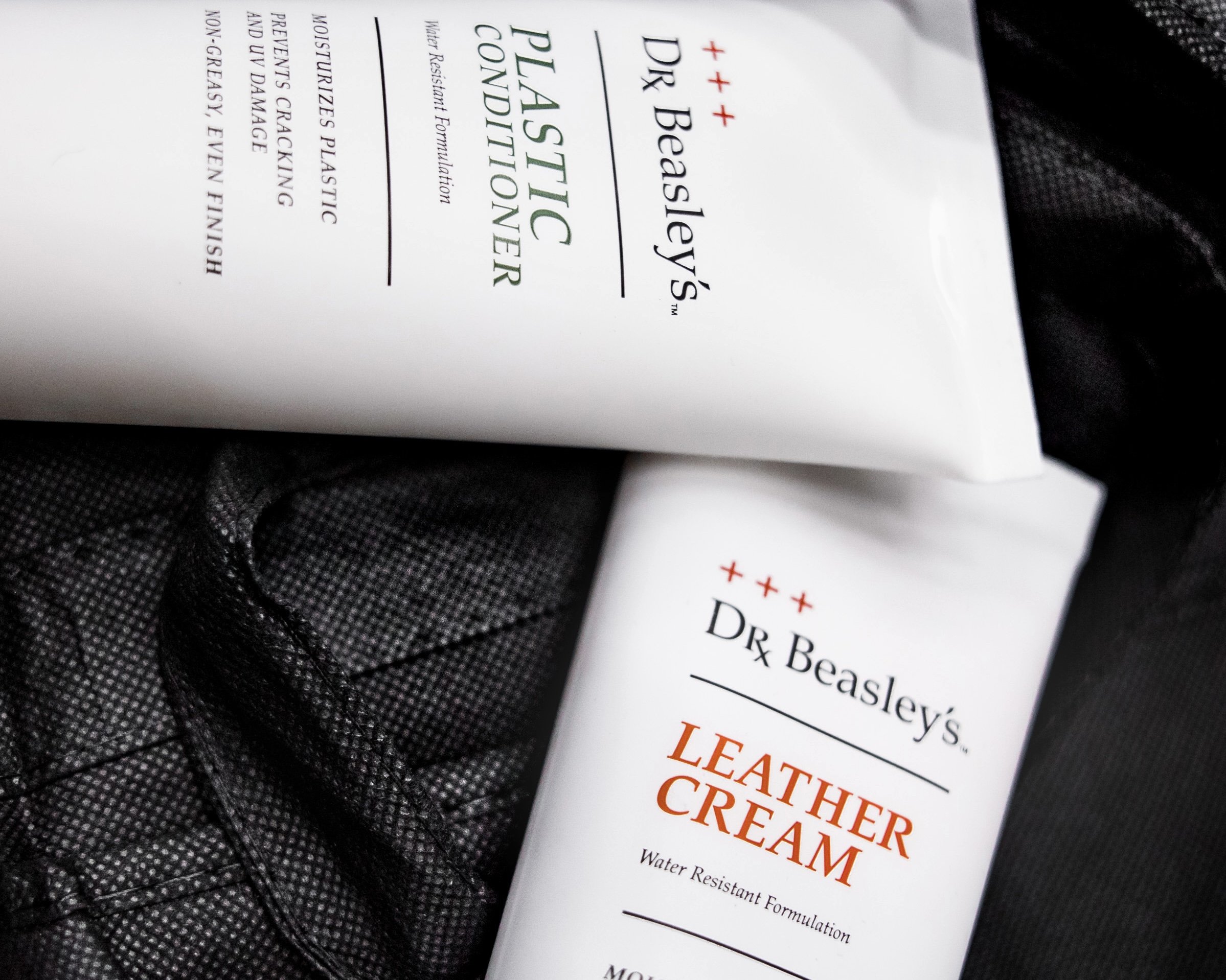 Two white tubes of Dr. Beasley's, a brand who carries detailing products in the car industry. The bottles are labeled Leather Cream and Plastic Coating, with black textured fabric in the background.