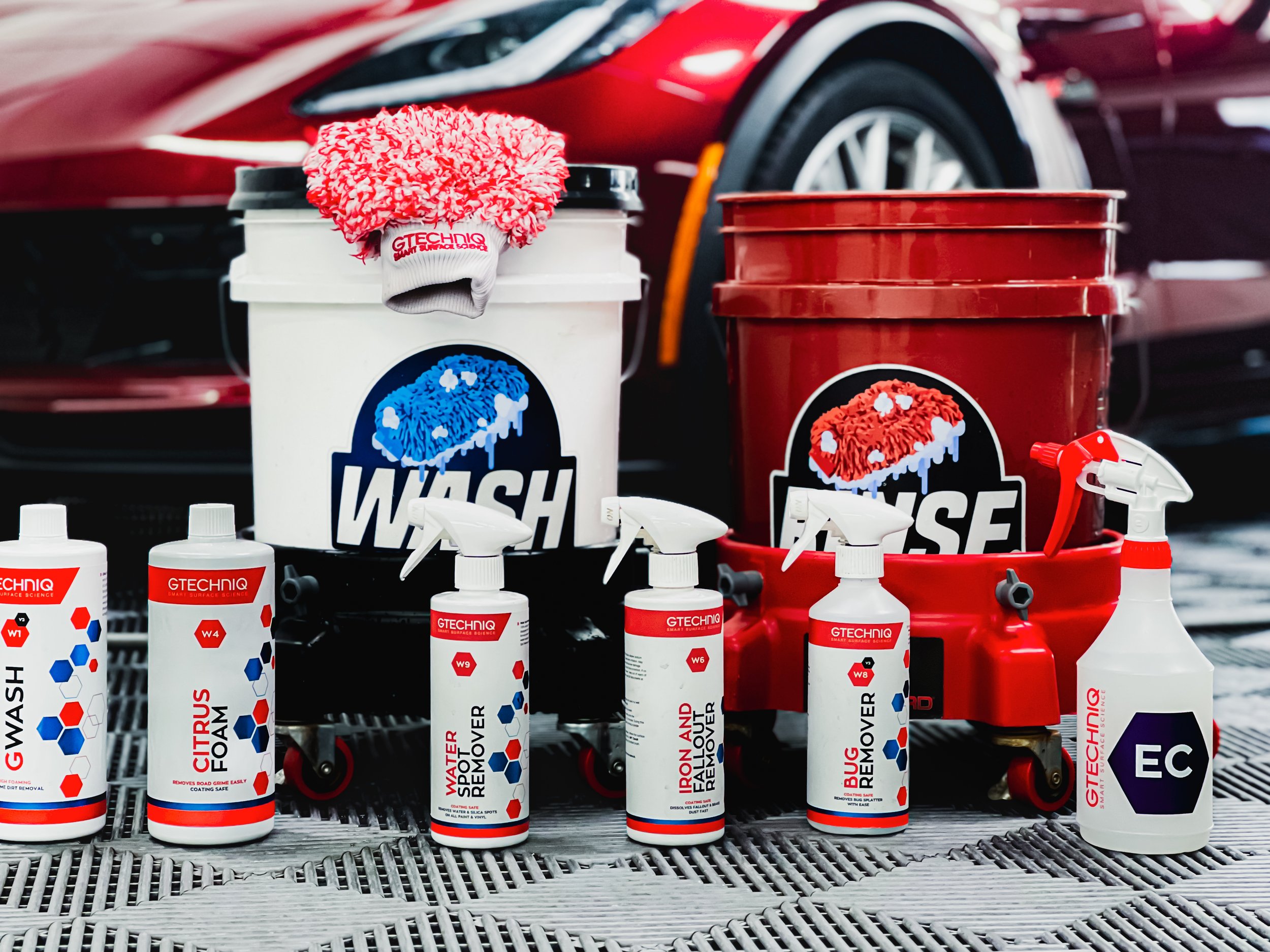 Car cleaning supplies including spray bottles, brushes, and buckets with the brand GTECHNIQ who is based in the UK with a North America headquarters. They are one of the leading detailing product brands in the industry. There is a red sports car.