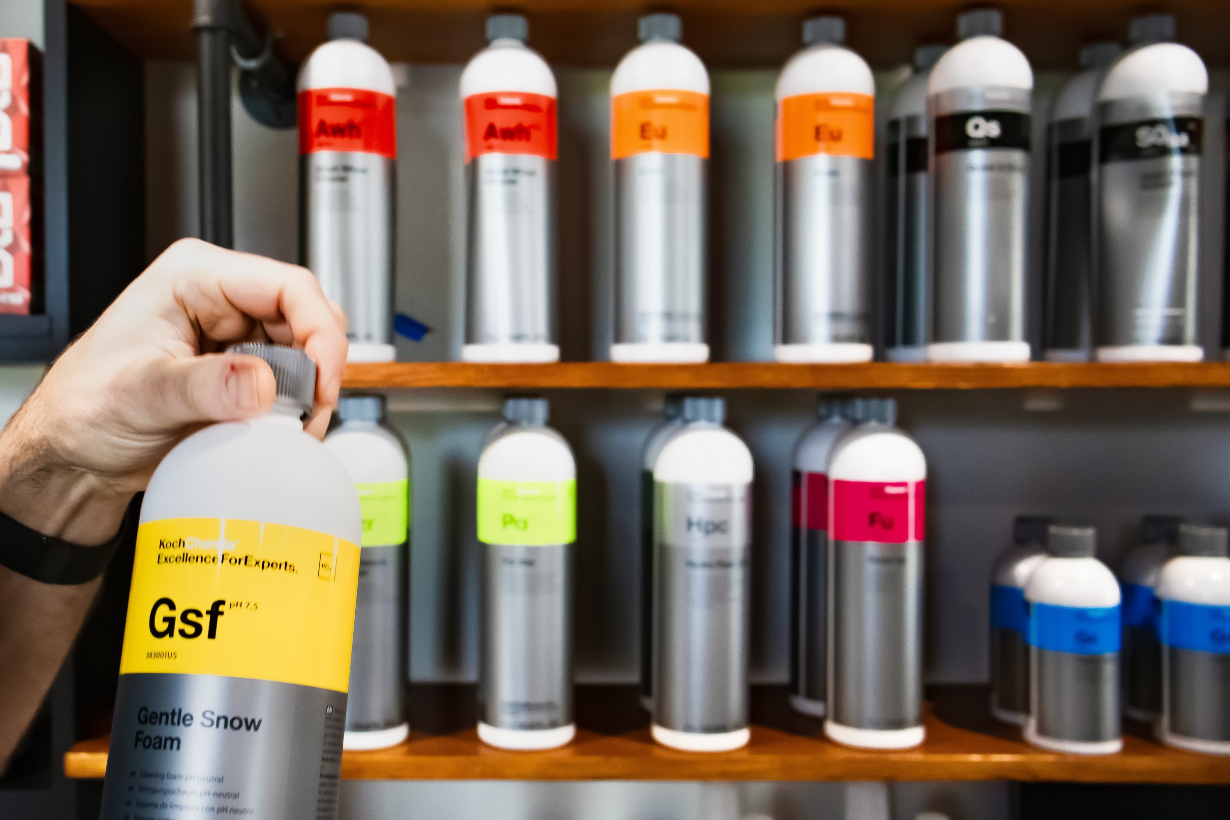 Person holding a bottle labeled 'Gsf' by Koch Chemie, with a background of shelves in the bays physical retail store. The detailing products are in various colors and labels. The colors are familiar colors to the car detailing community.