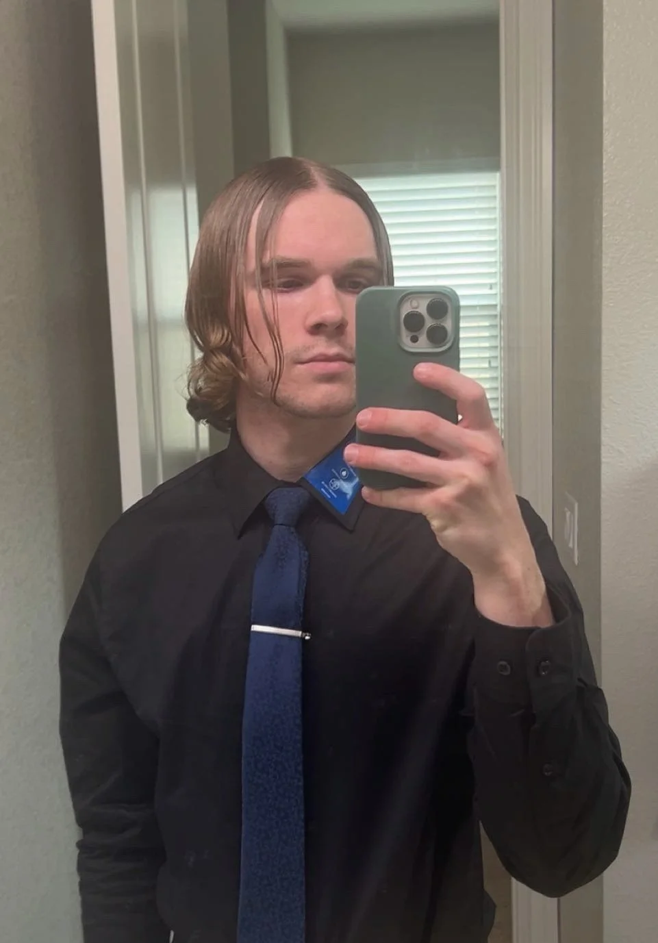 A person taking a mirror selfie with a smartphone. They have shoulder-length brown hair, wearing a black shirt with a blue tie, standing in a room with a window with blinds behind them.