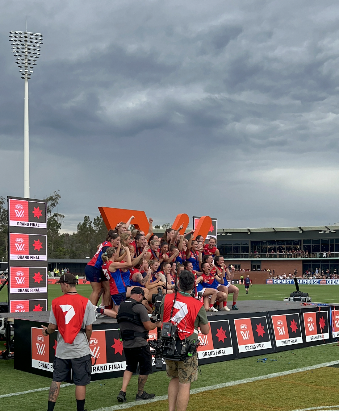 Australian football team celebrating on a podium after winning the grand final, with stormy clouds overhead and photographers capturing the moment.