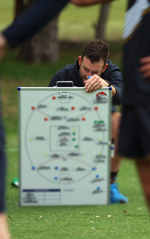 A man kneeling on the grass, resting his head on a clipboard with diagrams, next to a person in a suit, outdoors.