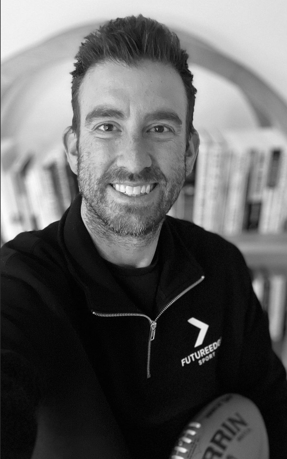 Black and white photo of a smiling man with short dark hair, a beard, wearing a black jacket with a logo that reads 'FUTUREEDGE Sport' and holding an AFL Ball in his lap, in front of a blurred bookshelf.