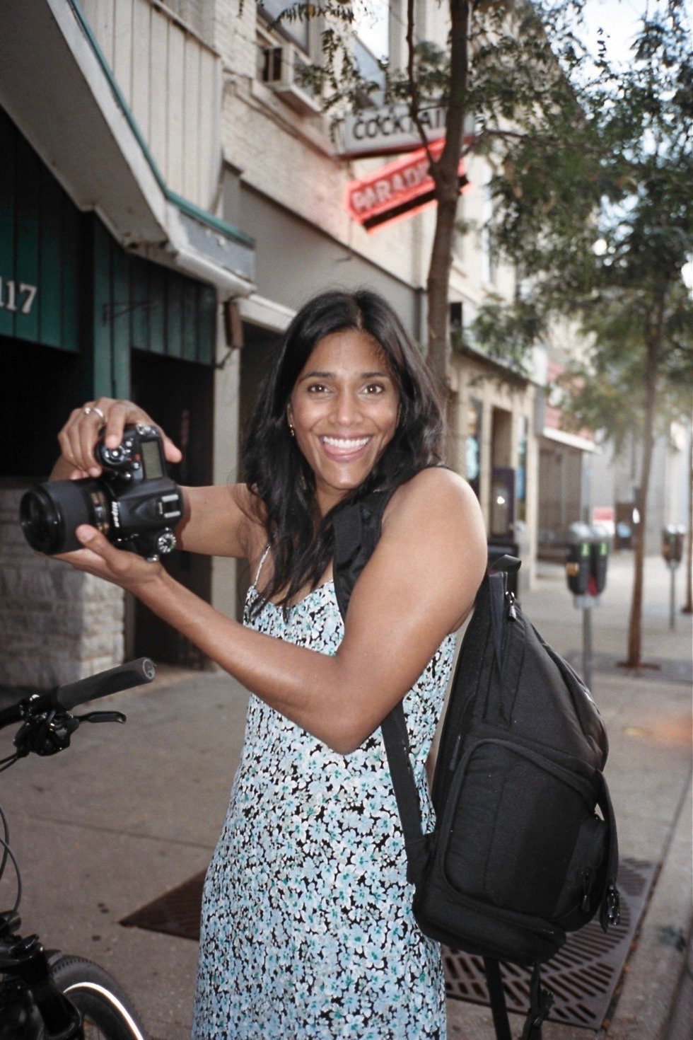 A smiling woman with long black hair, wearing a sleeveless floral dress and a black backpack, holding a camera on a city street with trees and buildings in the background.
