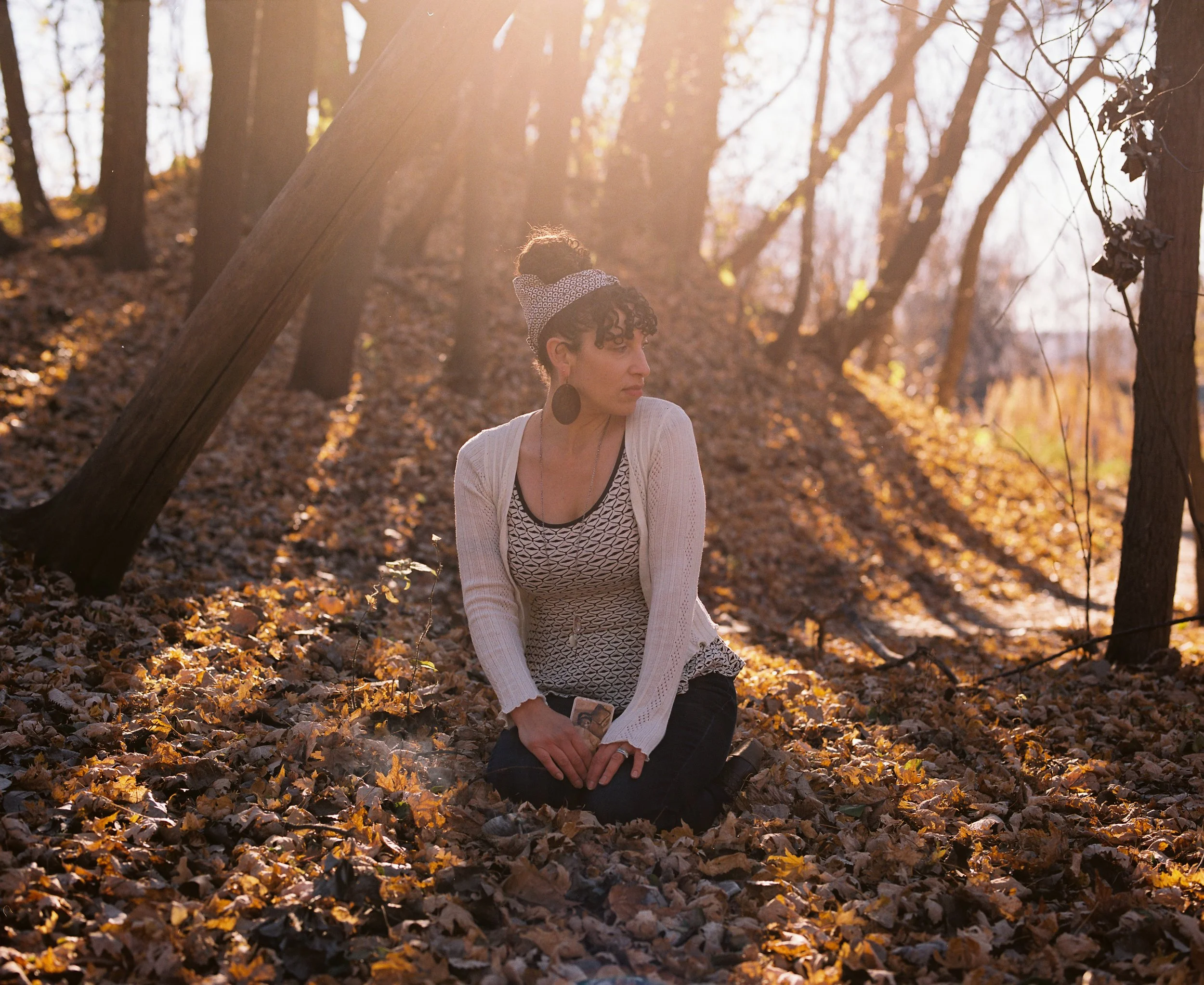 A woman kneeling on the ground in a forest during autumn, surrounded by fallen leaves, with sunlight filtering through trees.