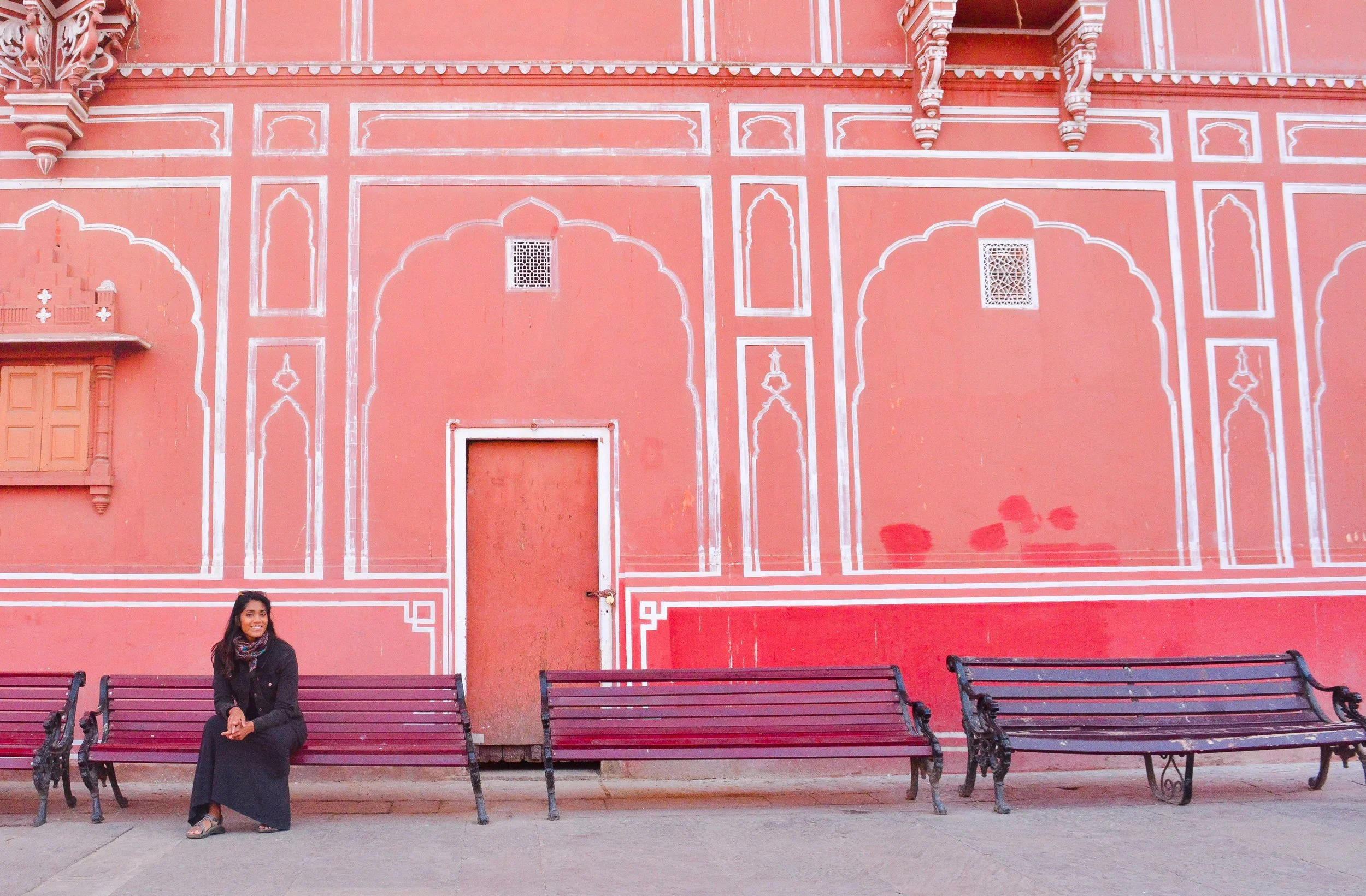 A woman sitting on a bench in front of a pink wall with white decorative lines and architectural details, with two empty benches beside her.